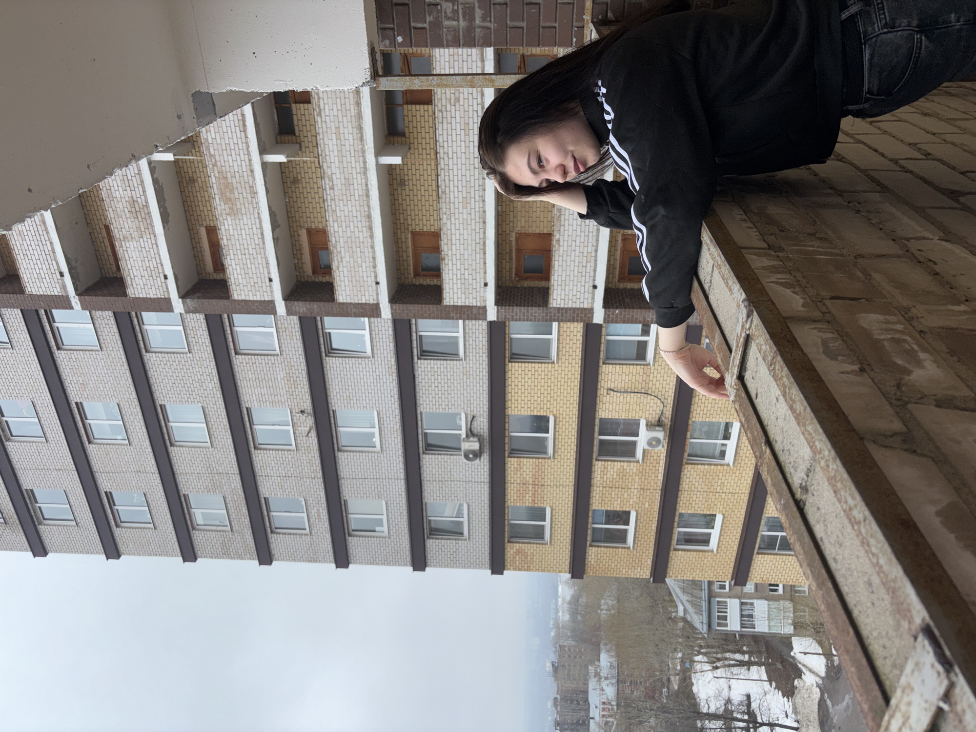 A young woman with long, dark hair is leaning on a brick ledge in front of a tall building. She is wearing a black jacket with white stripes on the sleeves and dark jeans. Her face is visible, and she appears to be looking downward. The ledge is made of light brown bricks and has a metal railing. The building behind her has alternating rows of light gray and yellow brick with windows. The location is likely in the city of Perm, Russia, given the urban architectural style. The sky is overcast. The camera is positioned at an unconventional angle, making the building and sky appear sideways. A section of a white concrete overhang is visible in the upper left corner. In the distance, houses and trees are visible beneath a cloudy sky.