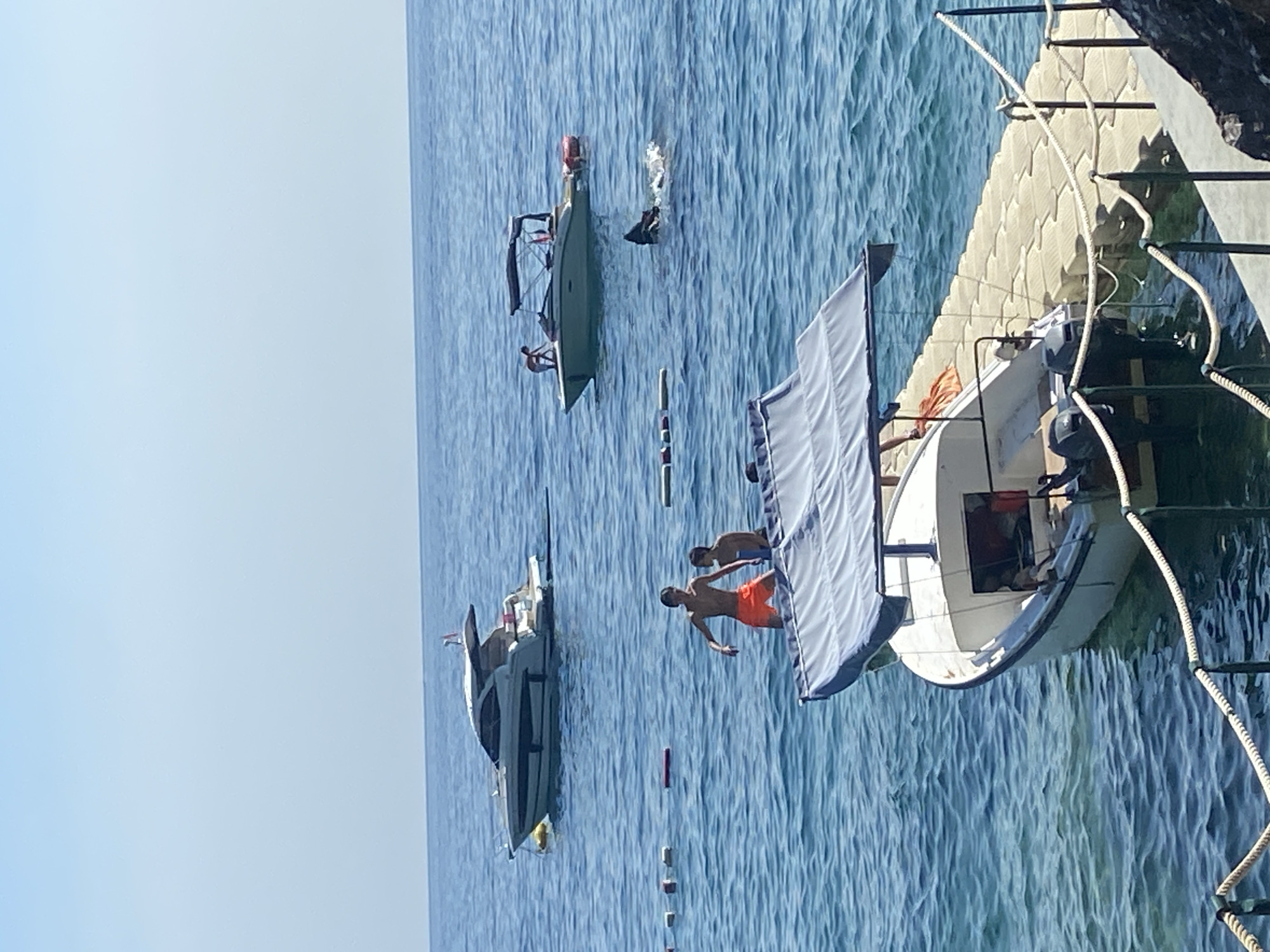 In Prijevor, Montenegro, on a clear day, a body of blue water is dotted with several boats and people. In the foreground, a small white boat with a blue canopy is docked at a floating pontoon. Two young men are standing in the water near the pontoon, one in orange shorts and the other in dark shorts. Further out in the water, a light-colored speedboat is anchored, with a person on board. Another, smaller boat is positioned in the mid-ground, with a person in the water next to it. In the background, a larger, white yacht is anchored. The sky is a pale blue, with no visible clouds. The water is calm, with gentle ripples. The scene appears to be a popular spot for recreational activities.