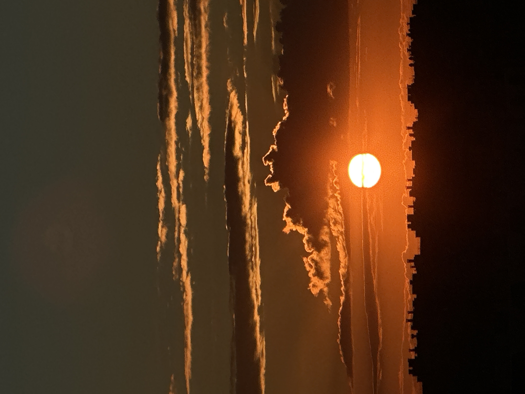 This is a photograph of a sunset, or sunrise, captured in a vertical orientation. The sky is ablaze with warm hues of orange and yellow, indicating the sun is low on the horizon. Wispy clouds are illuminated by the sun's rays, creating streaks of light that add depth and texture to the scene. The sun itself is a bright, glowing orb, partially obscured by the clouds.
On the right side of the frame, a silhouetted cityscape is visible, its jagged outline suggesting buildings and structures. This contrast between the vibrant sky and the dark urban silhouette creates a dramatic effect. The rest of the sky is a muted gray, fading to near black in the upper right corner, further emphasizing the brilliance of the sunset. The overall mood is one of quiet beauty and natural spectacle. The time of day is clearly either sunrise or sunset due to the sun's position and the intense colors in the sky. No people or specific objects are discernible other than the clouds, sun, and the distant cityscape. There is no visible text in the image.