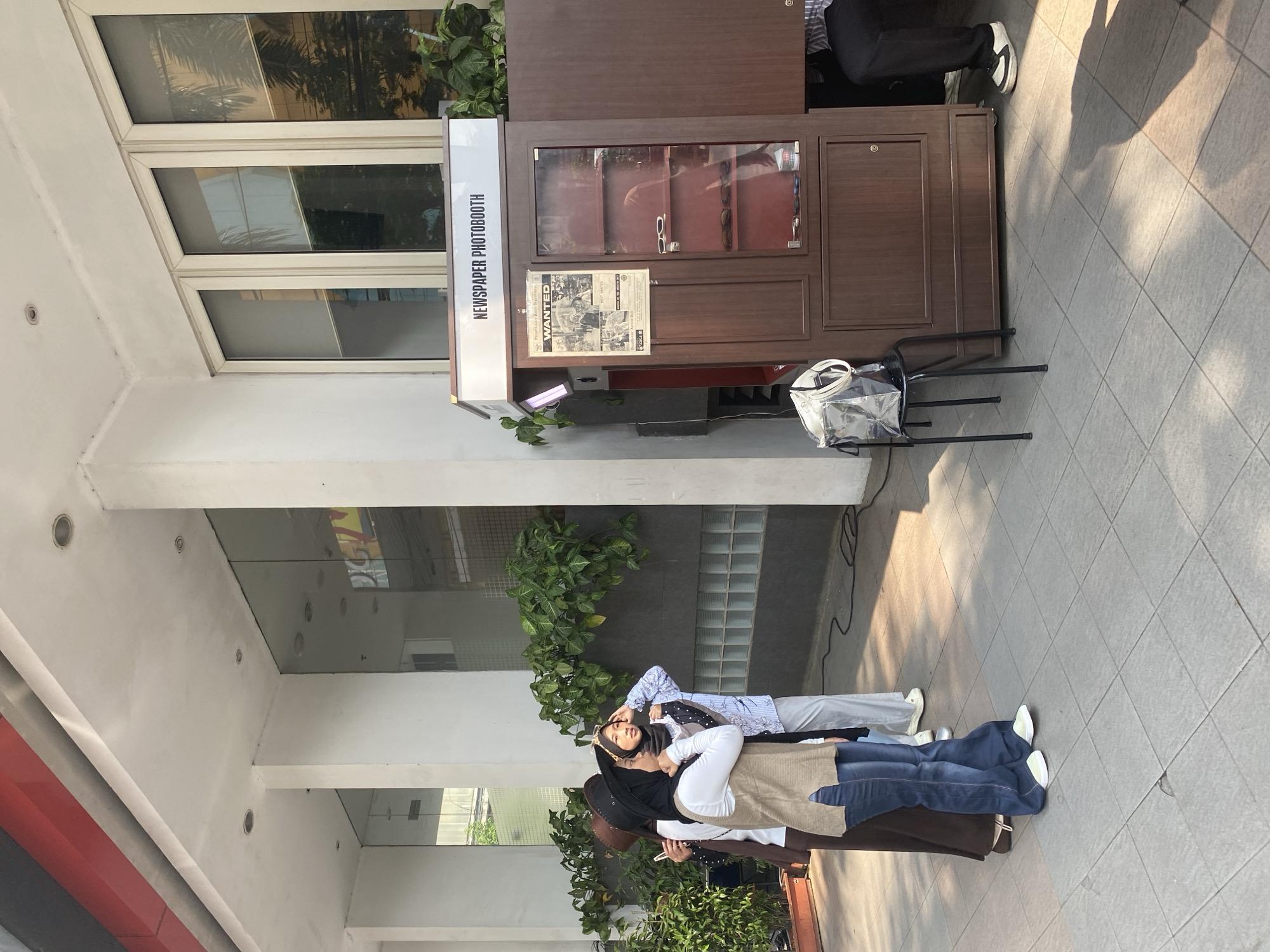 The image captures an outdoor scene in front of a building in South Jakarta, Indonesia. A "NEWSPAPER PHOTOBOOTH" is prominently displayed, a wooden booth with glass-fronted display cases showcasing sunglasses and other items, and a "WANTED" poster on its side. To the left of the photobooth, three young women are posing and interacting. One woman, wearing a brown hat and a white top with a beige vest and blue jeans, stands with her hand near her face. Next to her, another woman in a black hijab and a light blue patterned shirt, with a gold crown headband, also touches her face. A third woman, mostly out of frame, is also visible. A black metal stool with a transparent bag on it sits in front of the photobooth. The environment consists of a paved walkway with tiled flooring, a white building facade with large glass windows, and lush green plants. Shadows of people and objects are cast on the ground, indicating sunlight. The scene appears to be a casual outdoor gathering or event.