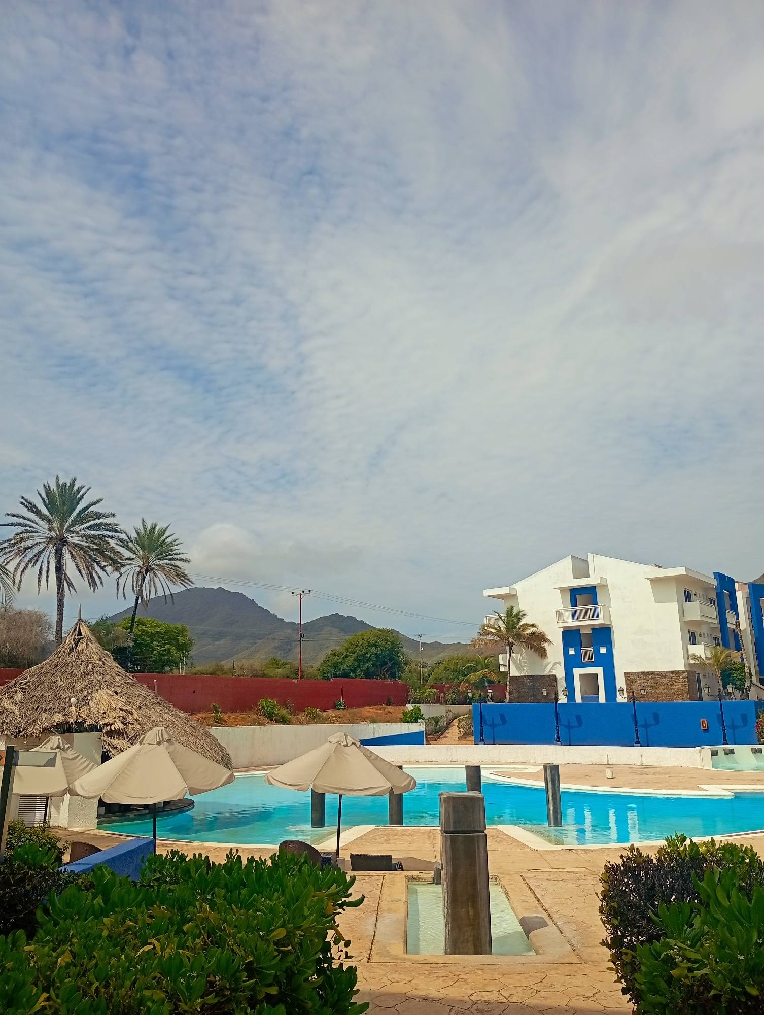The image depicts a resort scene in Playa El Agua, Venezuela. A large, light blue swimming pool with a connecting shallow area is in the foreground. Three grey, concrete pillars stand in the water. Adjacent to the pool is a seating area with three light-colored umbrellas and a thatched-roof bar. Green shrubbery is visible at the bottom of the frame, adding to the tropical ambiance.
Behind the pool, a white building with blue accents rises in the background. Palm trees stand to the left of the building, near a red retaining wall. Further back, a mountain range is visible under a partly cloudy sky. Power lines can be seen running across the mountains. The overall impression is that of a sunny, relaxed vacation destination.