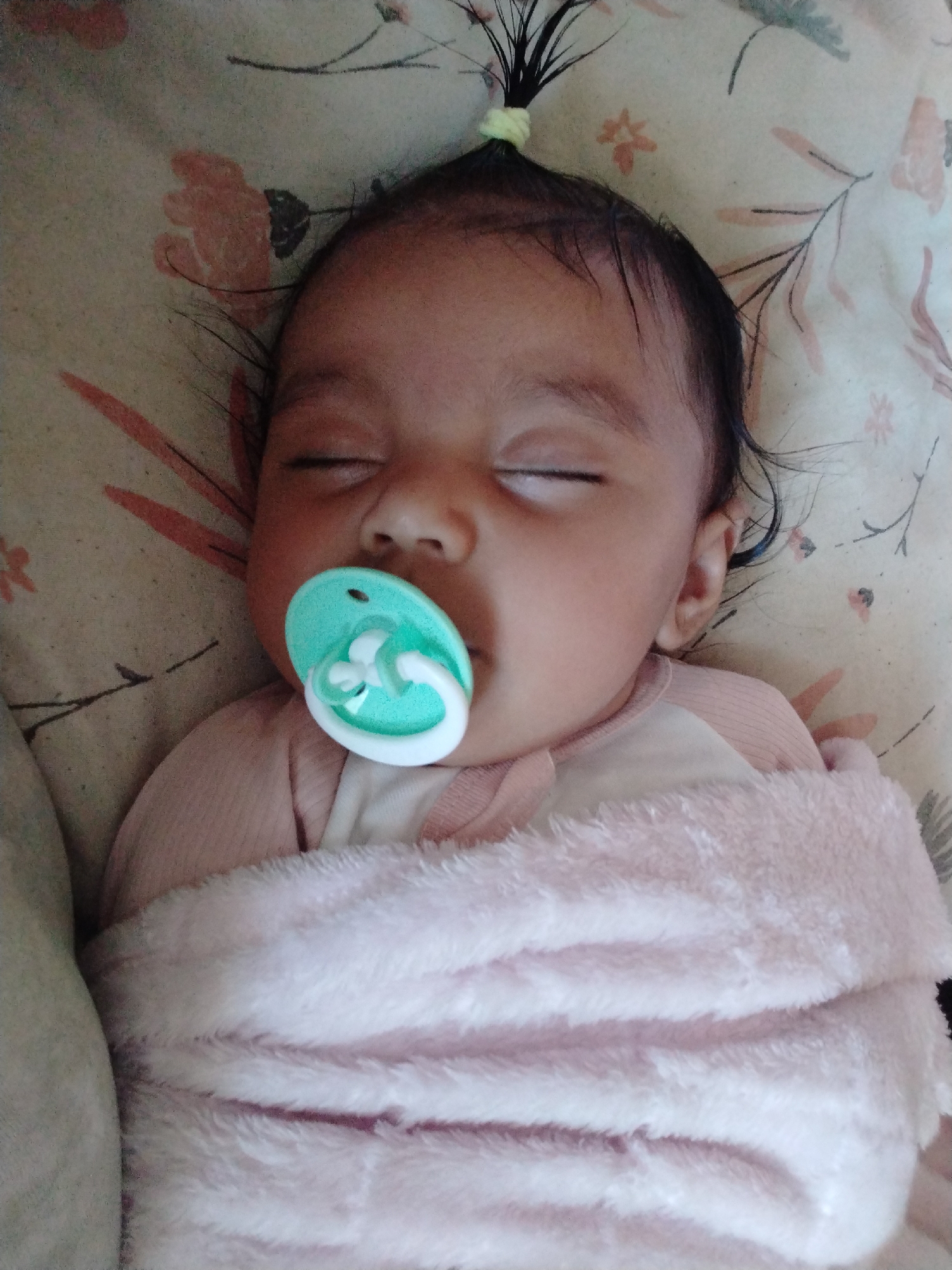 A baby with dark hair tied up in a small ponytail with a yellow hair tie is sleeping soundly with a turquoise pacifier in its mouth. The baby is wrapped in a pink, fluffy blanket and is lying on a bed with a floral pattern. The location is unknown.