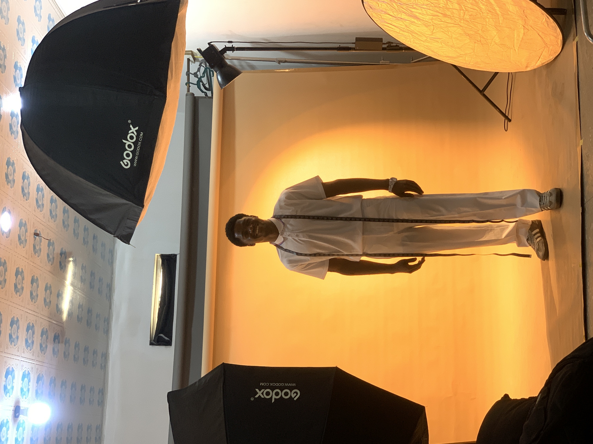 The image shows a man in white clothing, standing on a light orange backdrop, seemingly in a photo studio. The man is positioned horizontally in the frame and is wearing a white shirt and trousers with a dark measuring tape around his neck. He is also wearing a wristwatch. Two softboxes labeled "Godox" are visible on the upper left and lower parts of the image. There's also a reflector on the upper right. The background wall has light blue tiles with a floral pattern. There is an unidentified city, Gura Top, Nigeria indicated.