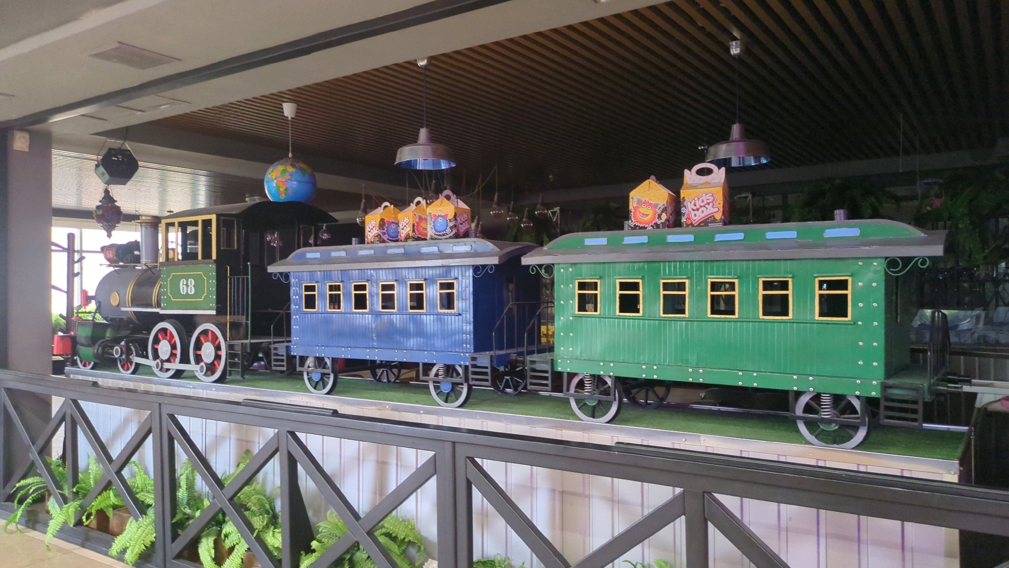 The image shows a decorative train display featuring a black locomotive with the number 68 on its side, followed by a blue passenger car and a green passenger car. Both passenger cars have multiple yellow-framed windows and sit on a grassy surface. Several colorful, box-shaped decorations are placed on top of the passenger cars, some featuring cartoon characters and text like "Kids Box." The train is positioned behind a dark grey metal railing with a criss-cross pattern. In the background, there are hanging lights and decorative elements, including a globe. The scene appears to be indoors, likely in a public space such as a shopping mall or entertainment venue. There are no people visible in the image. This scene is located in the city of Chișinău, Moldova, in the "Orășelul Copiilor" (Children's Town) amusement park located in the Râșcani district of Chișinău.