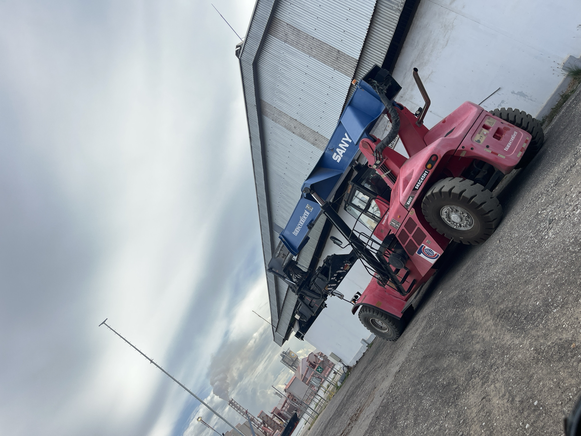 The image captures a red SANY brand reach stacker parked outside a corrugated metal building in Tambonbaroh, Indonesia. In the background, there are industrial structures and a communication tower, obscured by low-lying clouds. The sky is overcast. The ground is paved with a coarse, dark material.