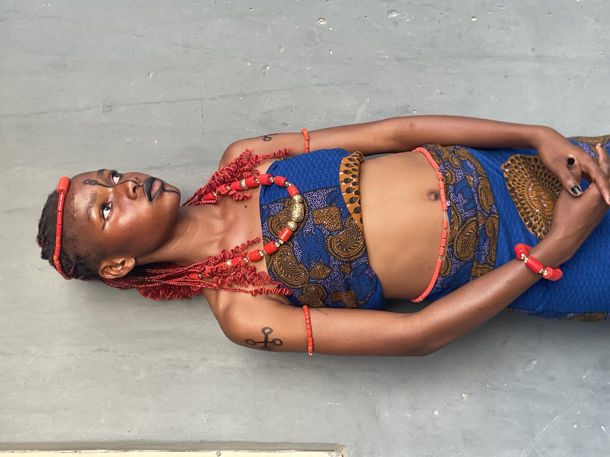 A young woman is depicted lying on her back on a plain grey surface, gazing upwards and slightly to her left. She is adorned in traditional attire, consisting of a vibrant blue and brown patterned fabric, possibly a wrapper or skirt and a matching strapless top.
Her ensemble is heavily accessorized with red coral beads, a significant element in many Nigerian cultures, particularly the Edo and Yoruba. She wears a multi-strand red beaded necklace featuring a prominent gold-colored centerpiece, red beaded bracelets on both wrists, and a single strand of red waist beads around her midriff. Her hair is styled in red braids, complemented by a red beaded headpiece.
Her face features distinct black linear markings around her eyes and mouth, along with dark lipstick, suggesting traditional face painting. Her expression is serene and contemplative as she looks upwards. She has an ear piercing and black nail polish on her fingers. A black tattoo-like symbol, resembling a cross with circular extensions, is visible on her left bicep. The overall lighting is bright and even, indicating it is daytime. The grey surface she is lying on serves as a simple, uncluttered background, allowing the intricate details of her attire and adornments to stand out. There is no visible text in the image.