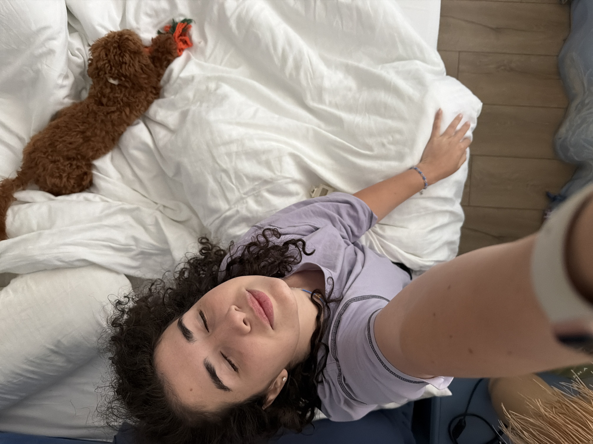 The image shows a woman lying on a white duvet with her eyes closed and a content expression. A small brown dog is lying on the duvet behind her, playing with a toy. The scene appears to be in a bedroom, possibly in the city of Banya, Bulgaria, given the context provided. The woman is wearing a light purple t-shirt. The room has wooden flooring and a blue headboard.