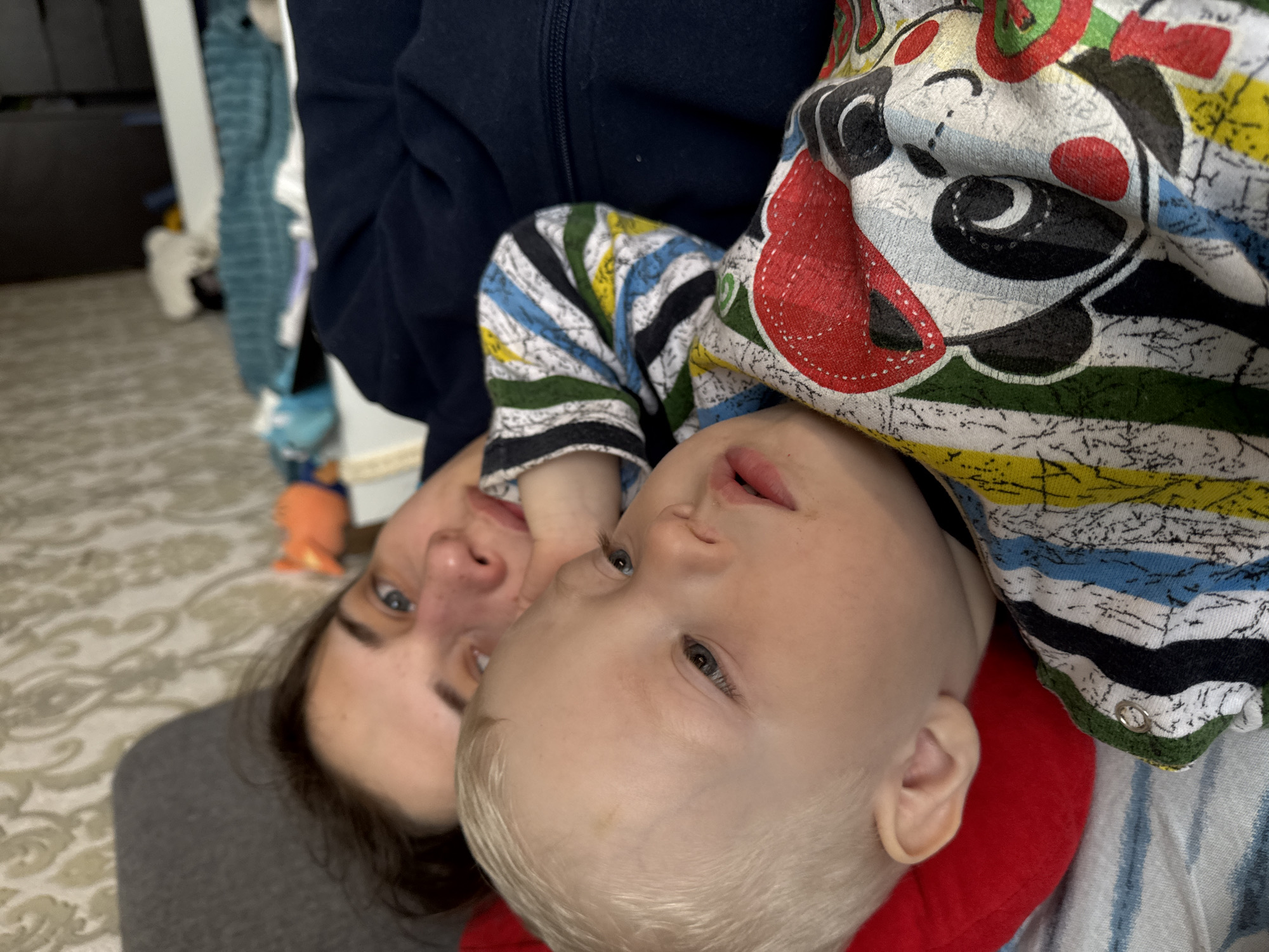 The image shows a woman and a baby indoors. The woman has dark hair and is wearing a grey shirt and a dark blue jacket. The baby has blond hair and is wearing a multicolored patterned outfit. They are positioned close to each other, with the baby lying on the woman's chest. The background includes a patterned carpet and a glimpse of some furniture. There is no information available to determine the location as Chaikovsky, Russia.