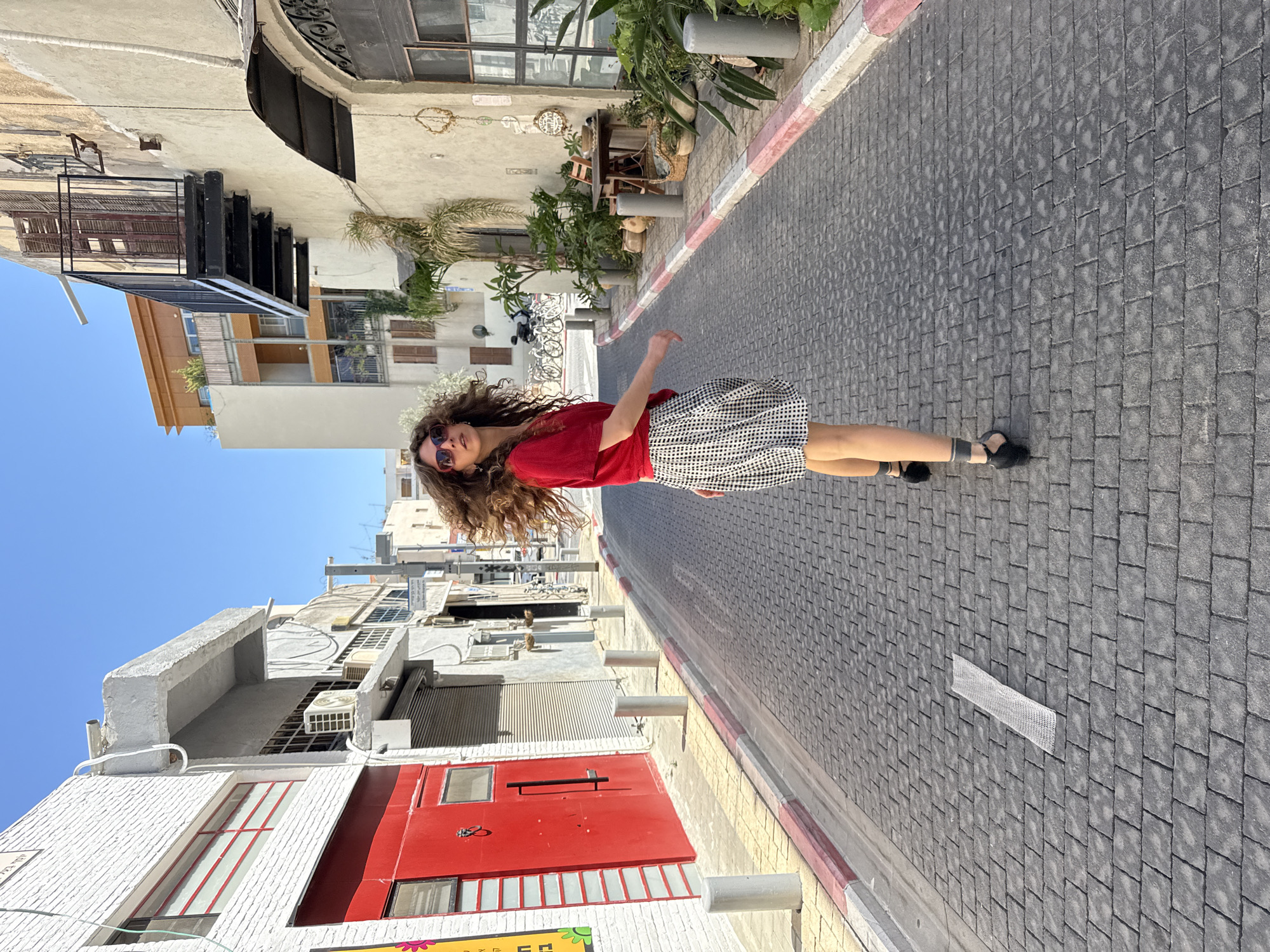 A woman with long, curly brown hair and sunglasses is walking down a narrow street in Tel-Aviv, Israel. She is wearing a red t-shirt and a black and white checkered skirt. Her hair is blowing in the wind as she walks away from the camera. The street is paved with cobblestones and lined with buildings on both sides. The buildings are a mix of modern and older architecture. On the left, there is a white building with a large red door. On the right, there are buildings with balconies and potted plants. The sky is clear and blue. There are several bicycles parked on the right side of the street.