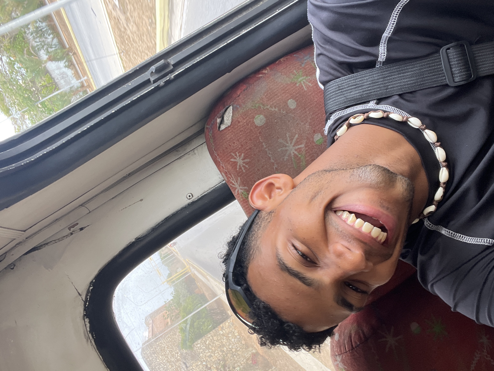 A young man with short, curly black hair is smiling broadly inside a vehicle, possibly a bus or taxi, as suggested by the worn red patterned seat. He is wearing a black athletic shirt with white stitching, a black belt, and a shell necklace. Sunglasses are perched on his head. He is looking directly at the camera. Through the vehicle's window, a blurred street scene is visible, indicating an urban environment. The location is likely Porlamar, Venezuela.
