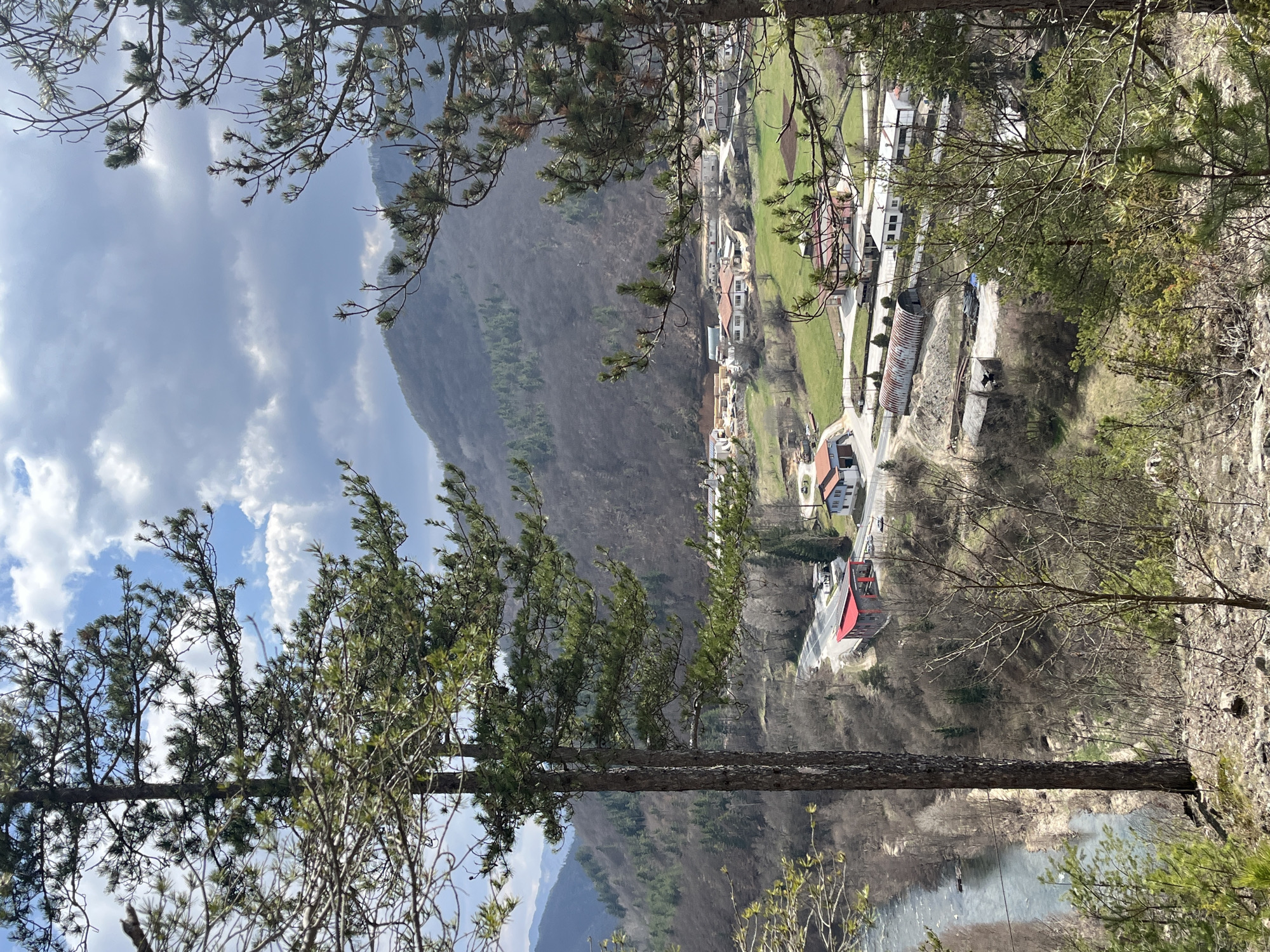 The image presents an elevated panoramic view of a valley in Srednogortsi, Bulgaria. The sky is partly cloudy, displaying both bright blue areas with white cumulus clouds and darker grey cloud formations. In the foreground, several pine trees with dark trunks and green needles, alongside bare deciduous branches, frame the scene from a rocky, sparsely vegetated slope.
A light blue-grey river is visible in the lower-left midground, flowing alongside a paved road. The road curves sharply to the right, revealing a modern-looking building with a prominent red roof and grey walls positioned adjacent to the curve. Further into the midground, a scattering of structures typical of a small settlement is present, including several white-walled houses with orange/red tiled roofs. An industrial area features a long, corrugated metal arch-shaped building and other smaller structures, with evidence of cleared land or construction nearby. Green grassy fields are interspersed between the buildings and industrial zones.
The background is dominated by forested mountains, appearing primarily deciduous with patches of conifers, suggesting a late winter or early spring season due to the bare tree branches on many slopes. No individuals or specific activities are discernible within the scene.