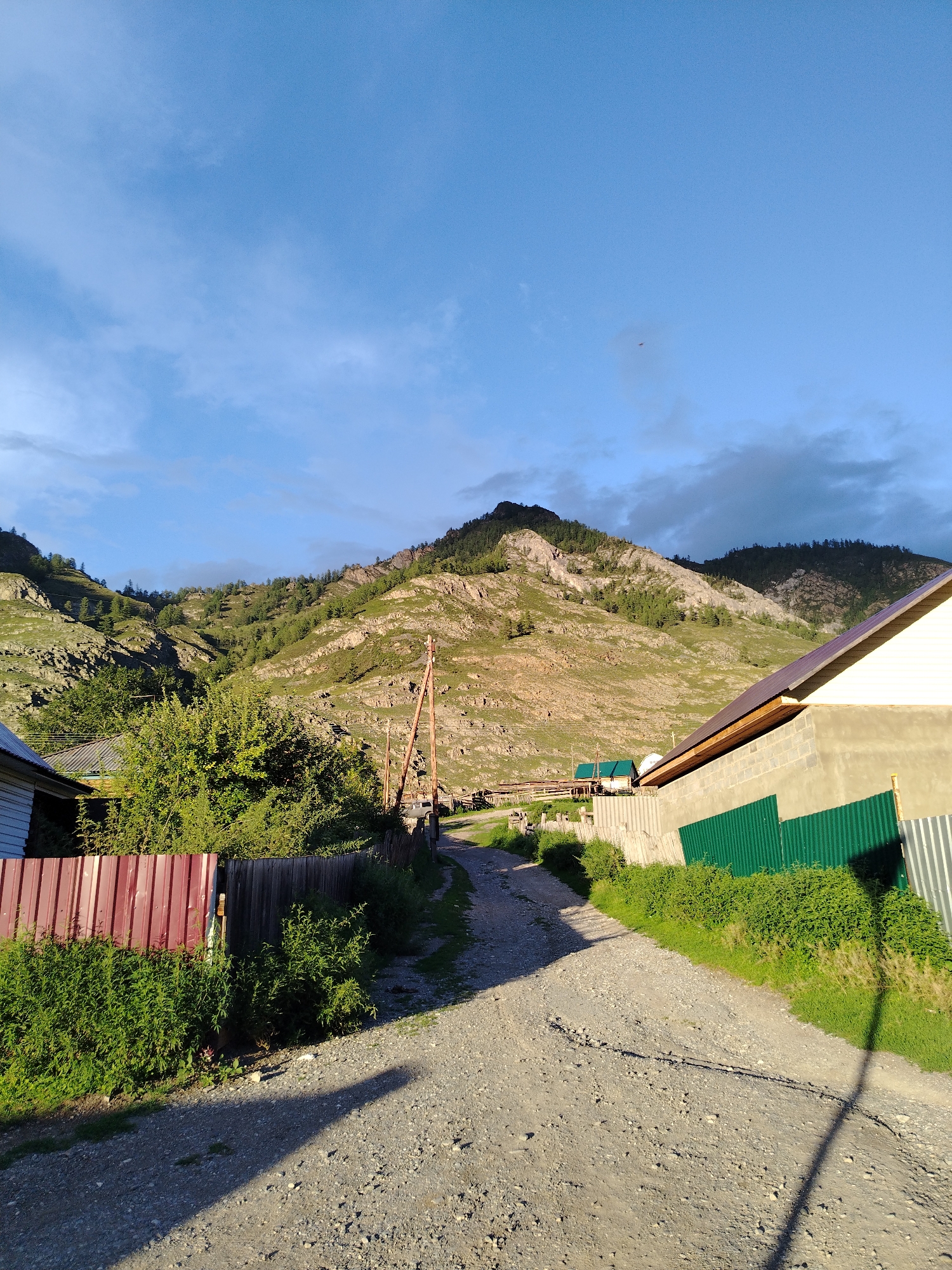 The image shows a rural scene in Iodro, Russia, with a dirt road leading towards a mountain range. On the left, a red metal fence encloses a yard with lush green bushes. A house with a white exterior is visible behind the fence. To the right of the road, a building with a light-colored exterior and a dark roof is situated, partially obscured by a green metal fence. The mountain in the background is rocky and covered with sparse green vegetation and trees. The sky is a clear blue with scattered clouds. A utility pole stands near the road. The overall impression is a serene, natural landscape with signs of human habitation.