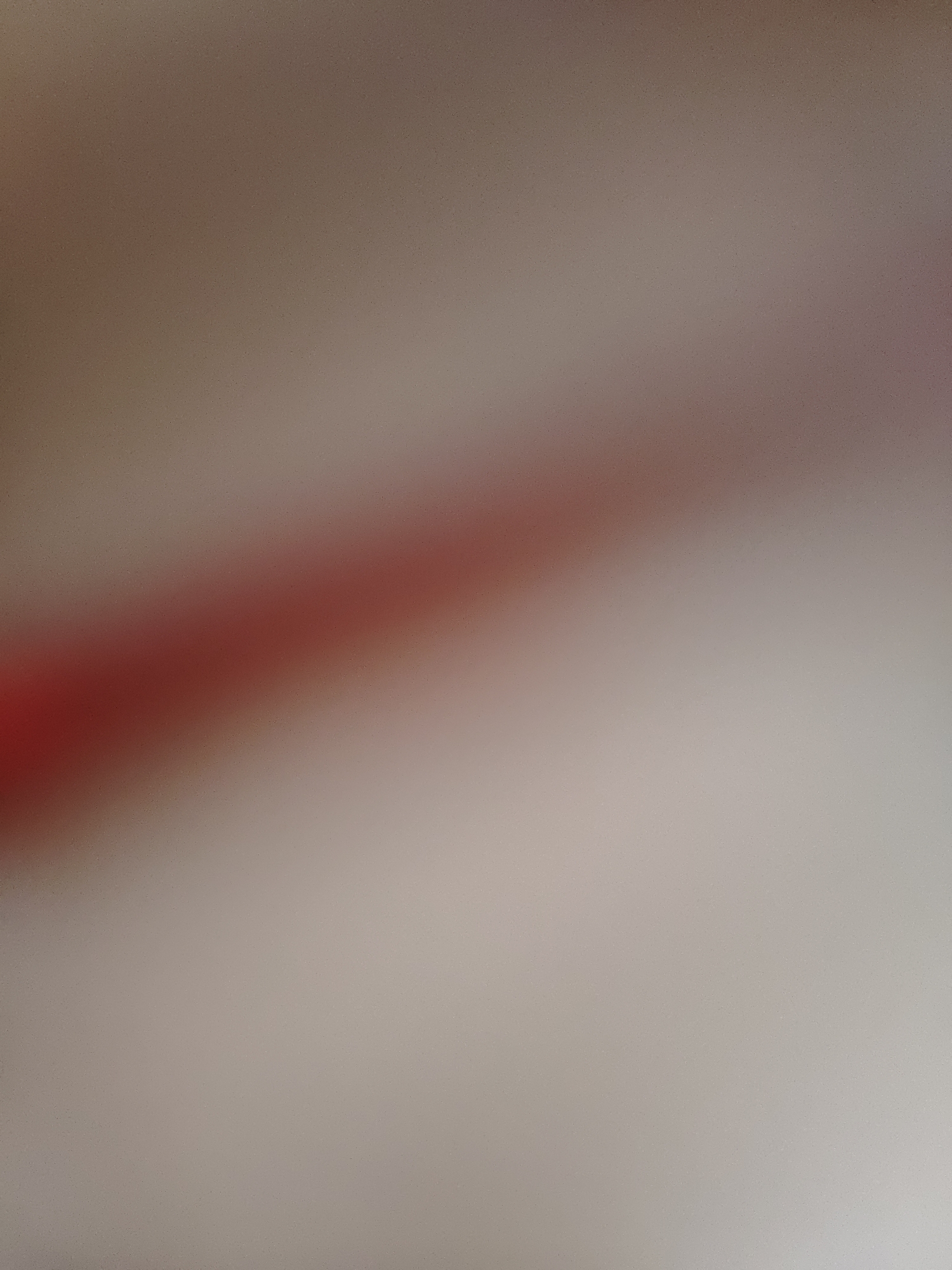 The image is an abstract, out-of-focus shot of a surface with a diagonal red stripe. The background is a gradient of muted browns and beiges, suggesting a textured material like fabric or paper. The red stripe is a vibrant, solid color that cuts across the image from the bottom left to the upper right. The edges of the stripe are soft and blurred, indicating the out-of-focus nature of the photograph. There is no discernible text, people, or any specific scene or event taking place. The lighting appears to be soft and diffused, with no harsh shadows. The overall impression is one of abstract color and form rather than a representational depiction.