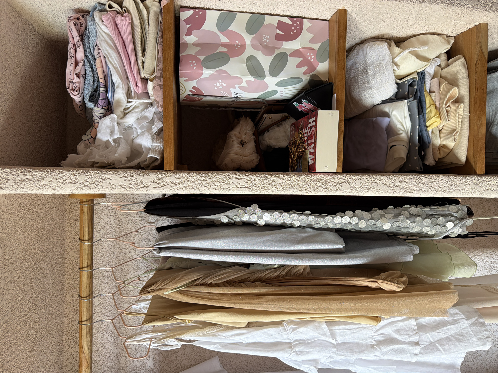 The image depicts a closet with three shelves and a hanging rod. The top left shelf contains folded clothing, including items in pink, gray, and white. The middle shelf holds a gift bag with a floral design, along with purses, wallets, and a book. The top right shelf contains more folded clothing. Below the top shelves is a wooden rod from which several items of clothing are hung on hangers, including garments in black, silver, gold, and white. The wall is textured. The location is Mengwi, Indonesia.
