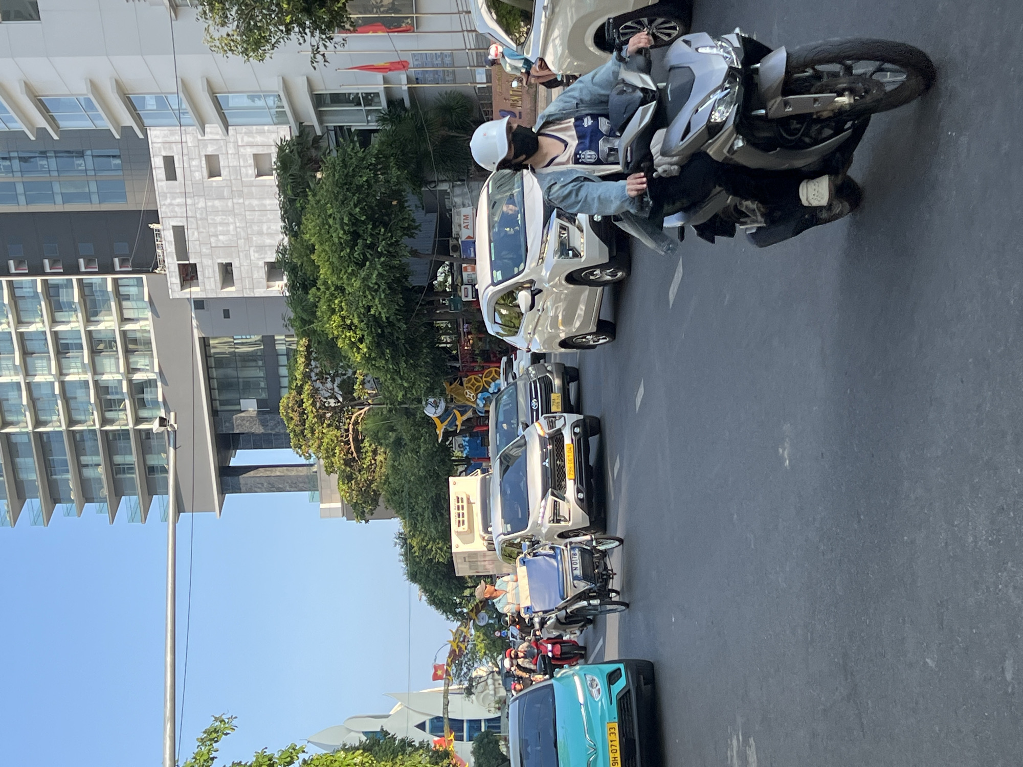 The image depicts a street scene in Nha Trang, Vietnam. The view is along a road with medium vehicle traffic, including cars and motorcycles. A person wearing a white helmet and a denim jacket rides a silver scooter in the foreground. Several cars are lined up along the road, with a blue car visible towards the end of the line. A building with a modern architectural style, featuring multiple floors and glass windows, is visible on the left side of the image. Trees line the street, partially obscuring other buildings further down the road. Vietnamese flags are also visible among the trees.