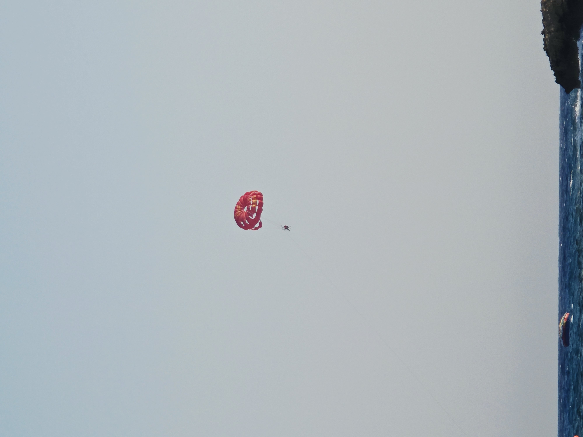 In the city of Ayia Napa, Cyprus, a parasailer is in the air, attached to a red parachute. They are positioned against a clear, light blue sky. Below, the deep blue sea is visible, with small waves. A rocky outcropping juts into the water on the right side of the image. There are a few buoys floating in the water.