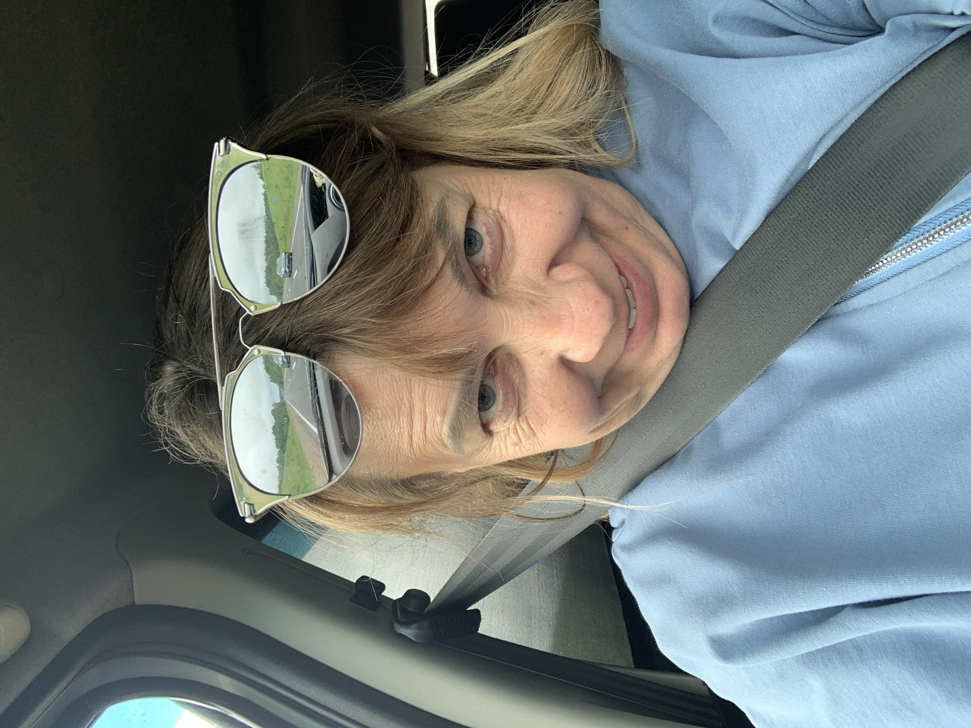 A woman wearing sunglasses is pictured from the chest up, inside a vehicle. She has blonde hair and is smiling. The sunglasses reflect a road with cars and greenery. The woman is wearing a light blue sweatshirt and a grey seatbelt is visible across her chest. The image appears to be taken in or around Хаценчыцкі сельскі Савет, Belarus, as indicated by the metadata.