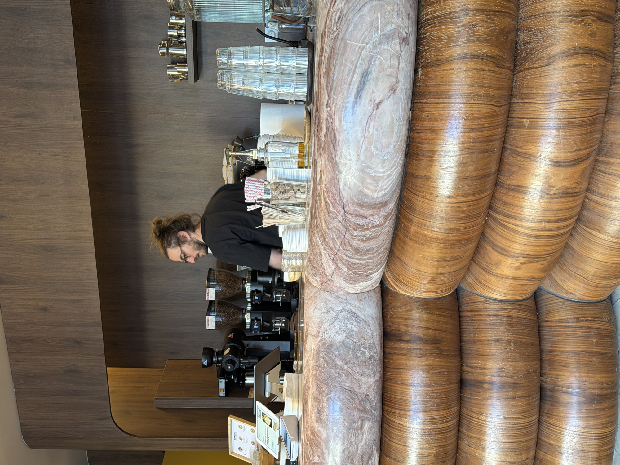 The image shows a man with a bun hairstyle standing behind a coffee bar, possibly in Paris, France. He is wearing a black shirt and appears to be working, perhaps as a barista. The bar is equipped with coffee grinders and other equipment needed for making coffee. The counter is made of marble, with wooden accents on its side. There are glasses and other containers visible on the bar. A digital tablet and a menu stand upright on the counter, near the coffee grinders.