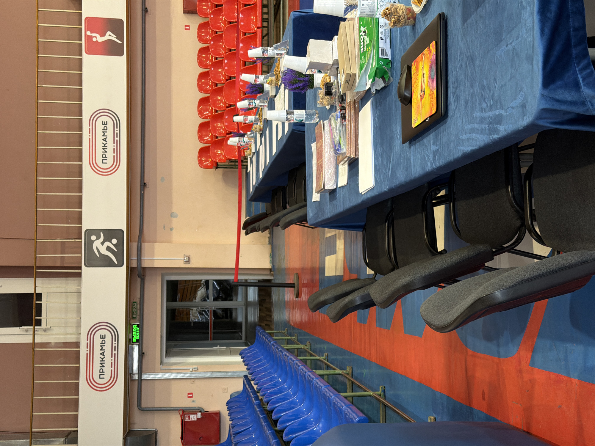 The image depicts the interior of the Molot Sports Palace in Perm, Russia. Rows of red and blue seating are visible on opposite sides of a multi-colored blue and red floor with chairs. Tables are draped with blue velvet-like cloth and are laden with various items: water bottles, cups, flowers, snacks, and documents. The tables are positioned in front of the seating. Vertical banners displaying the word "ПРИКАМЬЕ" (Prikamye) and pictograms of athletes are attached to columns. A green "EXIT" sign is visible above a doorway.