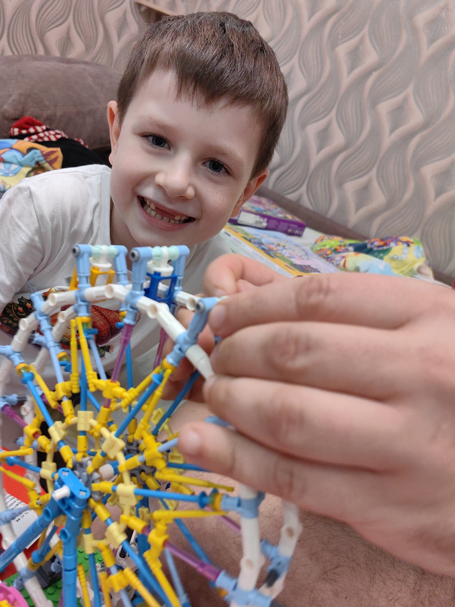 A young boy with brown hair and blue eyes smiles at the camera while a pair of adult hands builds a colorful construction toy in the foreground. The boy is wearing a white t-shirt and is positioned slightly to the left of the frame. The background consists of a textured wallpaper and a brown couch with some clothing items on it. The overall scene appears to be indoors. There is no information available to determine the location in the city of Sochi, Russia.