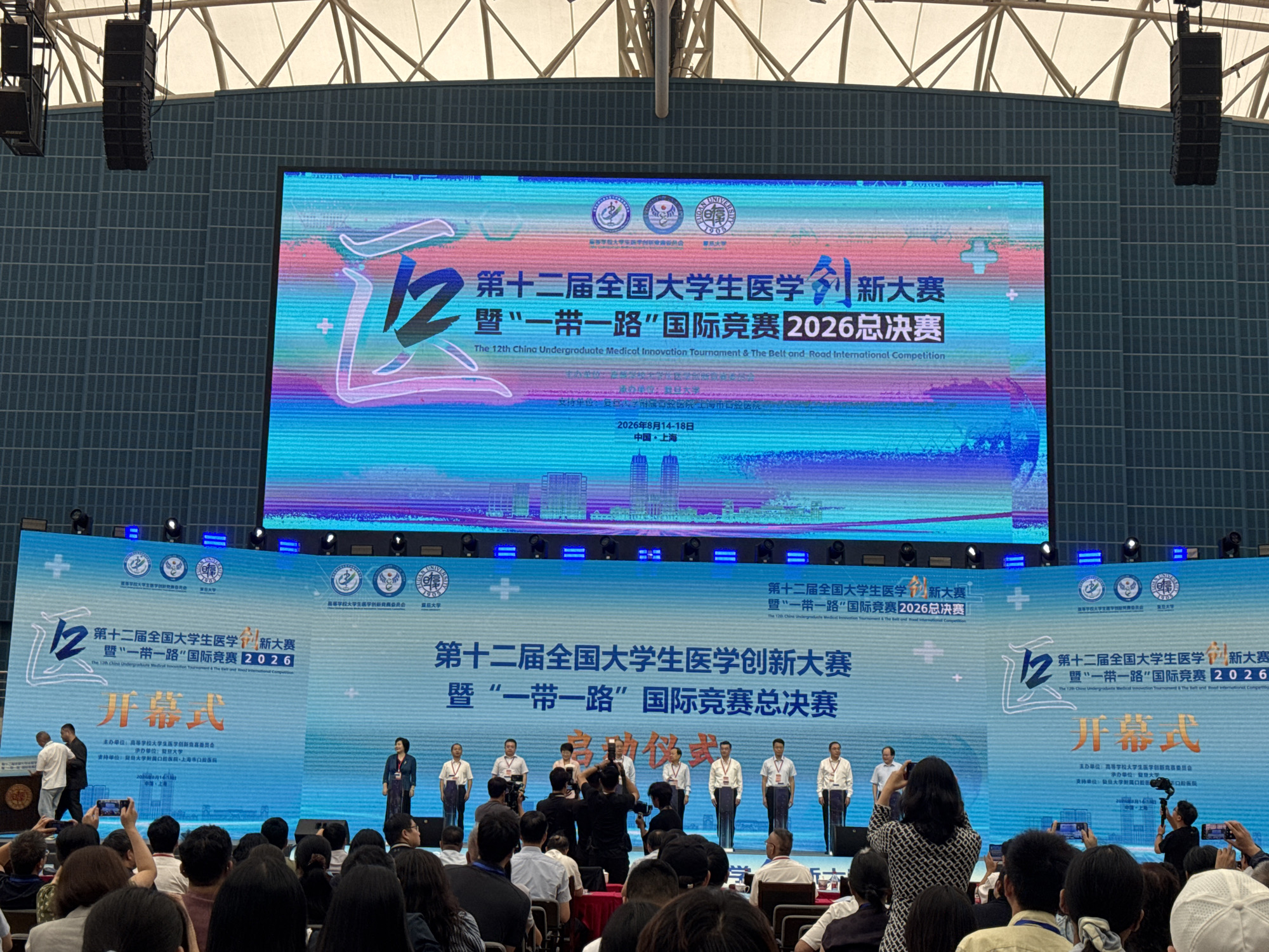 The image shows an indoor awards ceremony with a large LED screen displaying event information. The screen is in Chinese, with English text below it. The event appears to be the 12th China Undergraduate Medical Innovation Tournament and the Belt and Road International Competition, scheduled for 2026. The text on the screen announces "Opening Ceremony" in Chinese.
The scene is set in an auditorium or convention center, indicated by the tiered seating arrangement visible in the foreground, filled with a diverse audience. Several individuals, likely participants and organizers, are standing on a stage in front of the screen, some dressed formally. Two individuals are actively engaged in a handshake ceremony on the left side of the stage, while others stand in a line on the right. A photographer is visible in the audience, capturing the event. The background of the screen features a colorful, abstract design with city skyline elements. The ceiling of the venue is visible at the top, with structural elements and lighting fixtures.
The overall atmosphere appears to be one of formality and celebration, marking a significant event. The mention of "Yangpu District, China" in the prompt suggests the location is within that district, possibly at a university or conference venue.