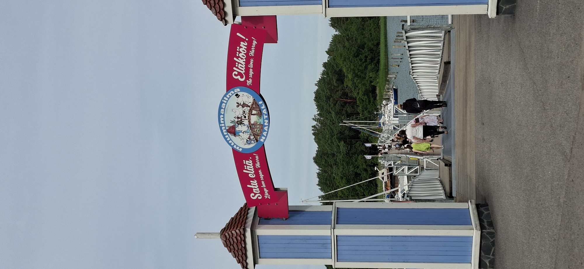 The image shows the entrance to Moomin World in Naantali, Finland. The entrance is a large, pink archway with text in Finnish and English, as well as a circular emblem featuring Moomin characters. To the left and right of the archway are blue and white towers with red roofs. A wooden pier extends from the entrance, with people walking on it. Boats are docked on either side of the pier, and a dense forest is visible in the background. The sky is overcast.