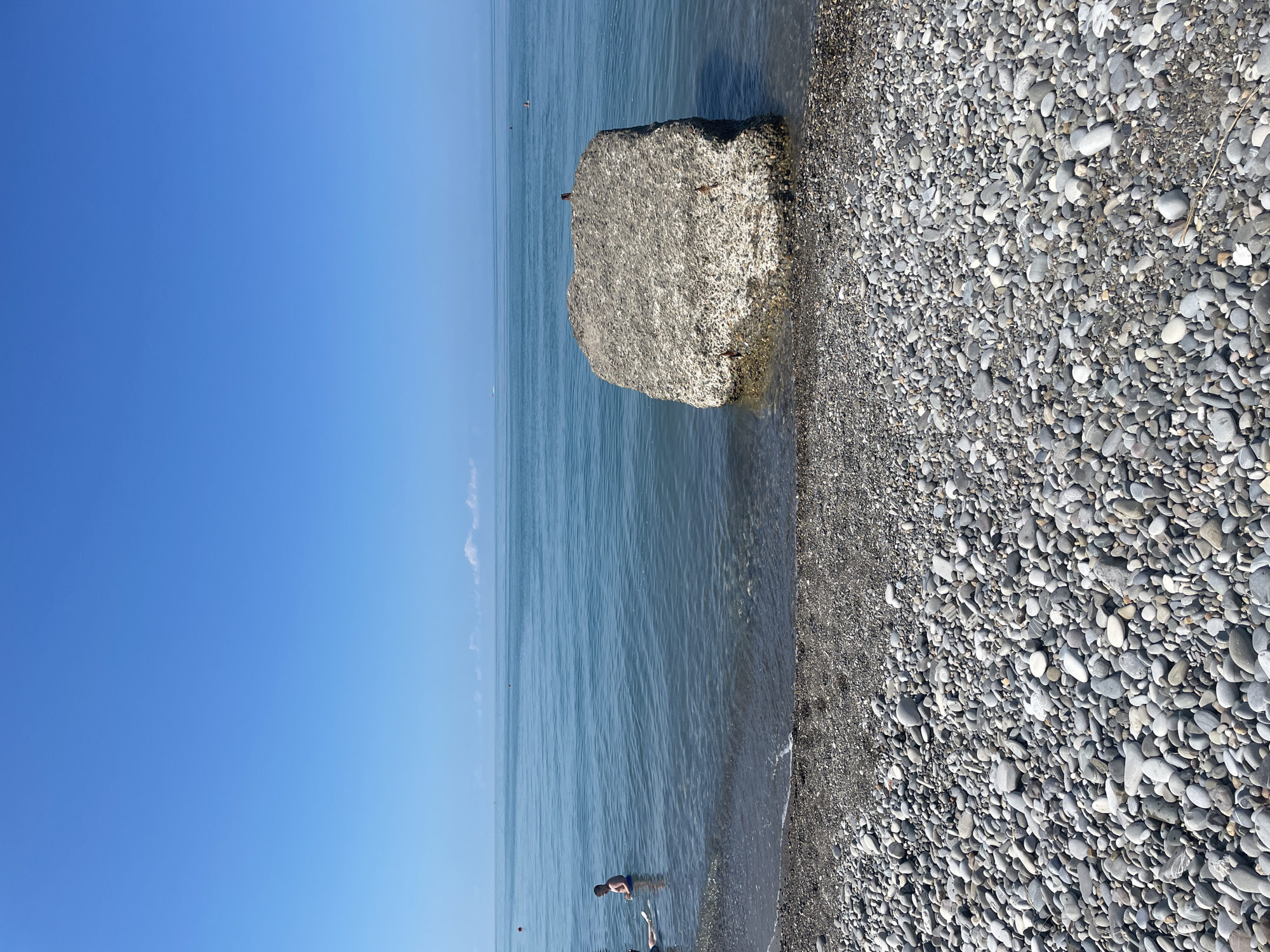 An eye-level, medium shot captures a tranquil beach scene under a clear blue sky. The foreground is dominated by a dense field of grey and white pebbles that form the beach. The middle ground features the calm, transparent blue water of the sea gently lapping at the pebbled shore. A large, rough concrete block sits partially submerged in the water, close to the shore. In the distance, the vast expanse of the sea meets the horizon under the unbroken blue sky. A few people are visible in the water: a person stands in the shallows on the left side of the frame, and another figure is seen further out in the sea. The overall impression is one of peacefulness and natural beauty.
