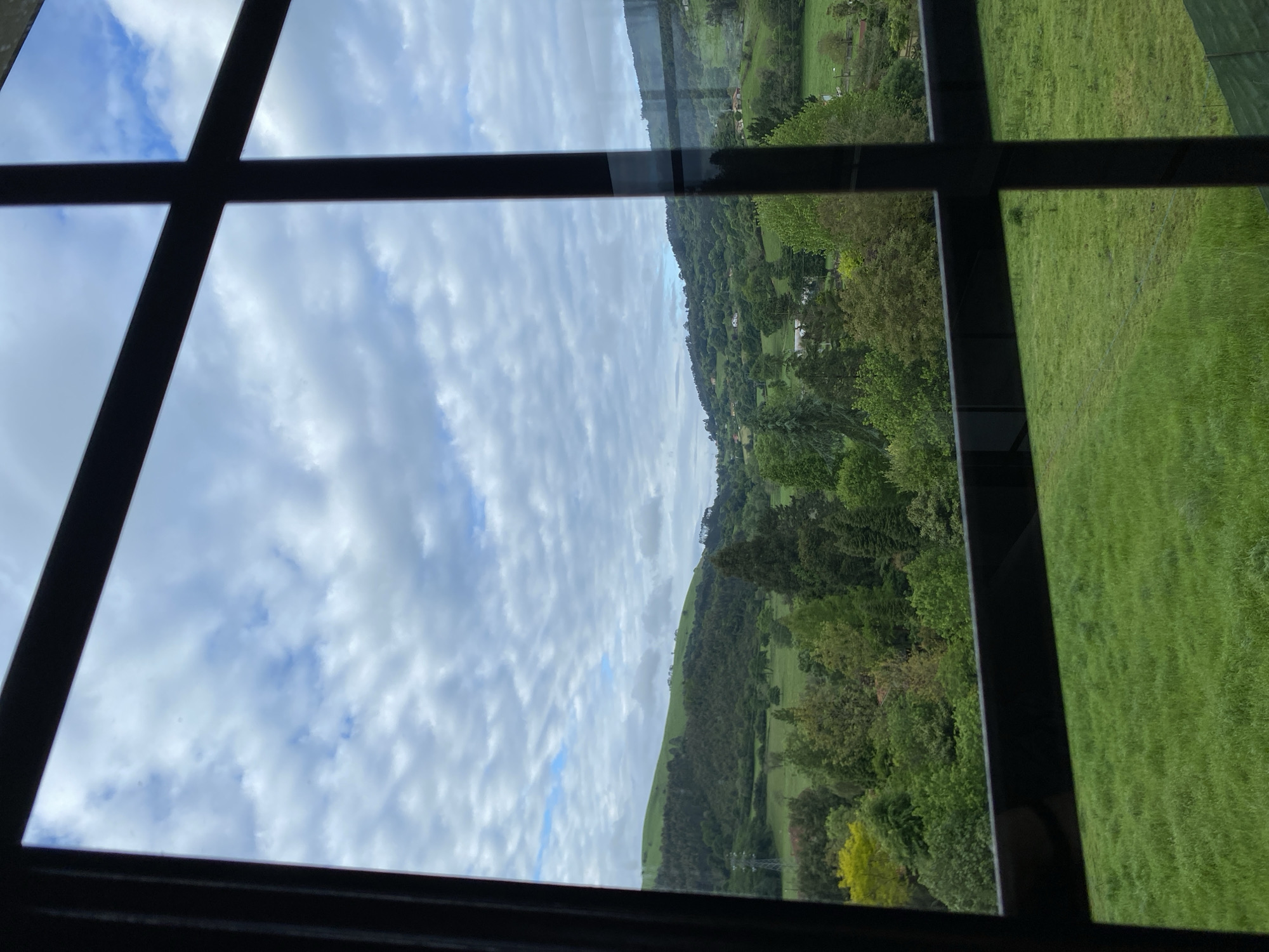 View through a multi-pane window showing a landscape. The landscape includes rolling hills covered in green grass and trees under a sky with scattered white clouds. The location is likely the countryside near Villaviciosa, Spain given the geography and vegetation. No people are visible.