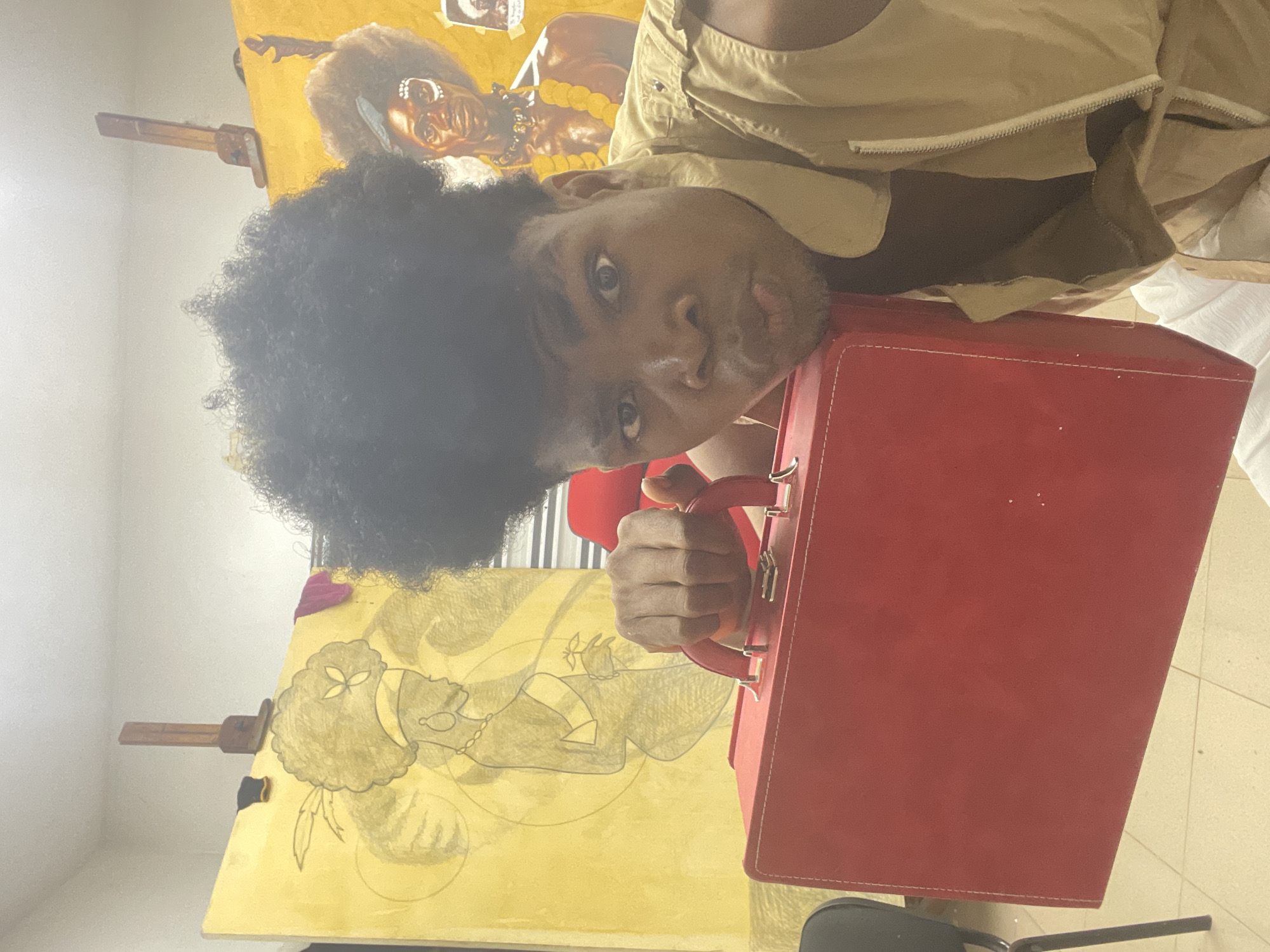 A medium shot captures a person with dark skin, curly black hair, and light-colored clothing posing with a red box. The location appears to be a studio in Ado, Nigeria. The walls are light-colored and adorned with large painted canvases featuring images of people. The foreground includes a black chair. The person is holding the red box with one hand.