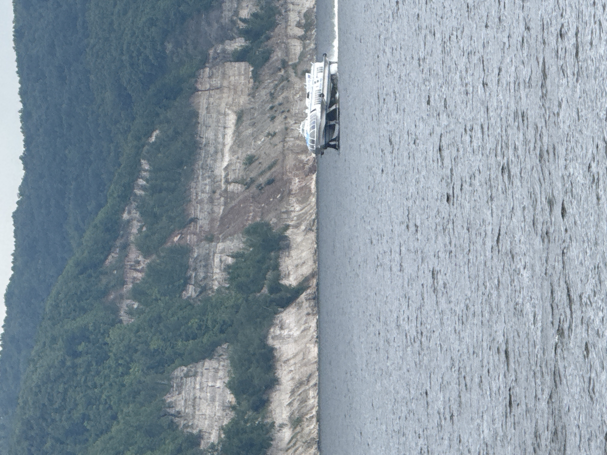 A white hydrofoil boat is moving from left to right on the Volga River in Kamskoye Ustye, Russia. The boat has a streamlined design with a glass-enclosed cabin. The river is calm with ripples on its surface. In the background, a steep, rocky cliff covered in green trees rises from the water. The sky is overcast.