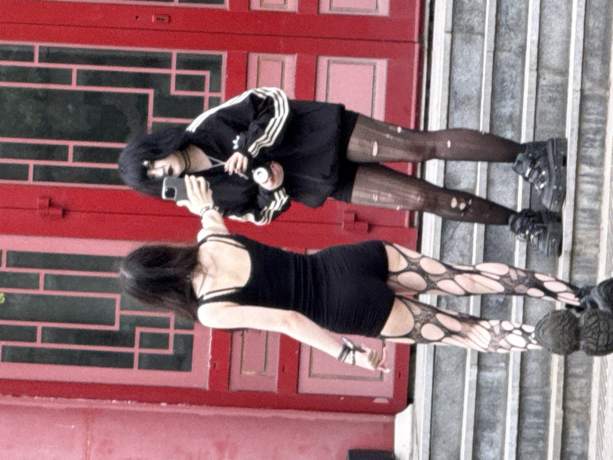 Two young women are standing on concrete stairs in front of a red building. One woman wears a black dress and torn tights; the other woman wears a black jacket with white stripes, a black skirt, and torn tights. The second woman is holding a cell phone and appears to be taking a picture of the first woman. The location is likely Moscow, Russia, based on the building architecture.