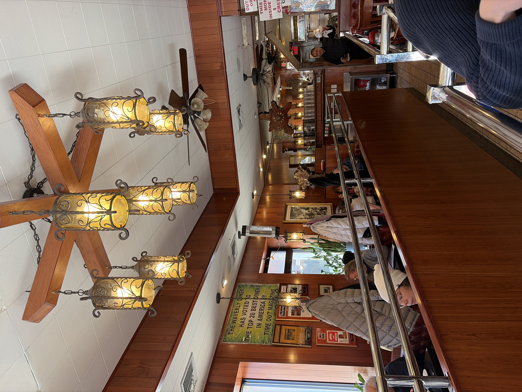 The image depicts the interior of the Waverly Diner, located in Greenwich Village, New York City, New York, United States of America.
The scene is an active diner environment, characterized by warm lighting and traditional decor. A prominent ceiling fixture crafted from wooden beams supports six ornate metal lanterns with amber glass, illuminating the foreground. The ceiling features white acoustic tiles, with several ceiling fans, some equipped with lights, visible throughout the space. Walls are primarily covered in reddish-brown wood paneling. A large green sign on the left wall proclaims, "THE WAVERLY DINER HAS VOTED TO THE TOP 20 BEST DINERS IN AMERICA BY TIME OUT MAGAZINE," flanked by framed accolades and other signs, including one that reads "Order up!".
Red upholstered booths are arranged, with a long, wooden partition topped with a polished chrome railing separating seating areas. Several patrons are visible; in the mid-ground, a person wearing a white knit beanie and eyeglasses is seated in a booth. Two quilted jackets, one grey and one white, are draped over booth backs. In the background, a male server, dressed in a dark shirt, stands near a service counter, holding a small white lidded dish in one hand and a dark object in the other. Adjacent to him is a small metal shelving unit. Indoor plants are situated near the windows on the left, offering a glimpse of the exterior. The floor is tiled.