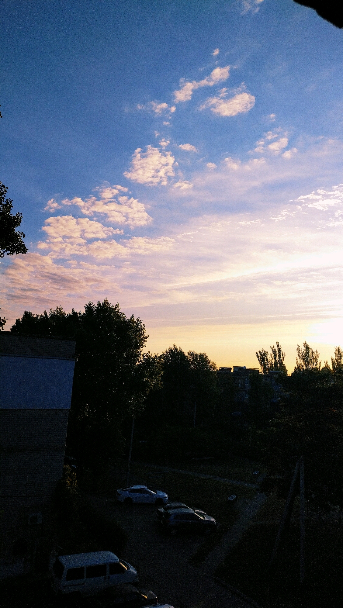 The image captures a dusk scene in the city of Horishni Plavni, Ukraine. A wide expanse of sky dominates the upper portion, featuring a gradient of blue near the top transitioning to pale yellow and pink hues near the horizon, indicative of sunset. Scattered white and pinkish clouds are visible throughout the sky.
Below the sky, a line of dark trees silhouettes against the lighter sky. In the foreground and midground, several cars are parked on what appears to be a residential street or courtyard. These include a white van, a white sedan, and a darker colored SUV. A portion of a brick building with a pale blue section is visible on the left side of the image. The overall atmosphere is quiet, with no people visible. The scene suggests a residential area within the city as daylight fades.