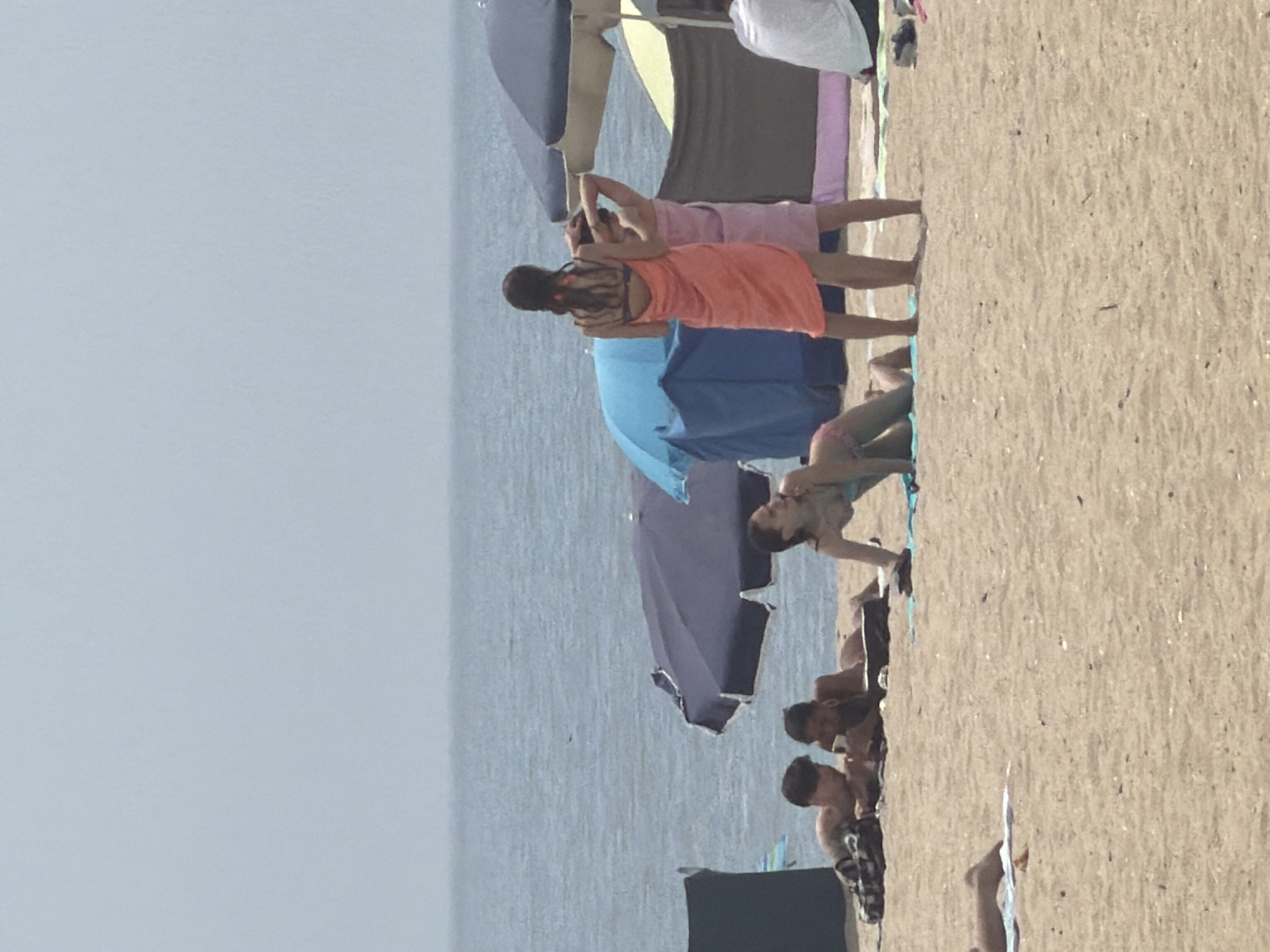The image depicts a beach scene with several individuals. In the foreground, a woman is positioned on her hands and knees on the sand, wearing a light-colored swimsuit. To her left, two men are lying on the sand, appearing to be looking at a mobile phone. Further to the left, a person's legs are visible lying on a towel. In the mid-ground, two women are standing, one with a dark braid is wearing an orange wrap, and the other, wearing a pink wrap, has her arms raised. Behind them, beach umbrellas are visible, one blue and one dark purple. The background consists of the sea and a hazy sky, suggesting a clear, overcast day. The location is the city of Espinho, Portugal, known for its sandy beaches and popular boardwalk.