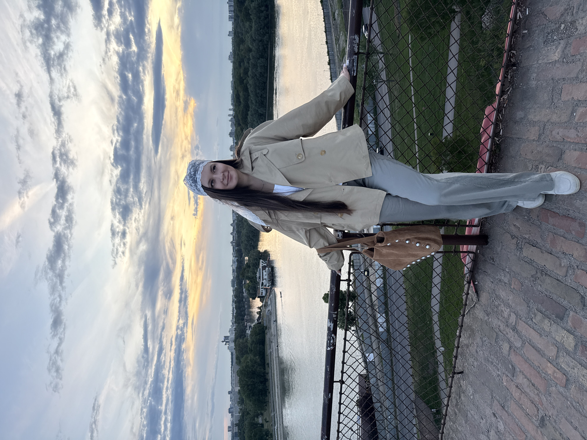 A full shot depicts a woman standing on a brick structure, leaning against a metal fence, with an urban landscape and a river visible in the background. The woman is dressed in light-colored clothing: a beige jacket, light blue trousers, and white shoes. She is wearing a white lace headscarf. A brown bag is hanging from the fence near her. The fence overlooks a river, with buildings and trees lining the opposite bank. The sky is partially cloudy, with some areas displaying a golden hue, suggesting either sunrise or sunset. Given the context and visual cues, this location appears to be a high vantage point, potentially part of the Stari Savski Most bridge in Belgrade, Serbia, known for its views of the city and Sava River.