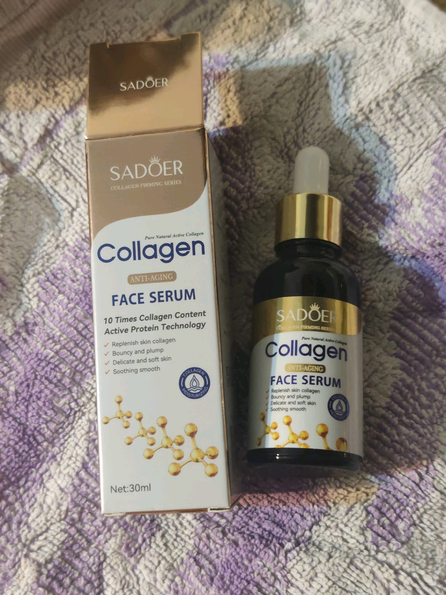 The image shows a Sadoer Collagen Face Serum bottle and its box on a textured, light-colored surface, possibly a blanket or towel. The box is open, revealing the product. The bottle is dark glass with a gold cap and a dropper. Both the bottle and the box have the brand "Sadoer" and "Collagen Face Serum" prominently displayed, along with text indicating "Anti-Aging" and "Pure Natural Active Collagen." The box also lists benefits such as "Replenish skin collagen," "Bouncy and plump," "Delicate and soft skin," and "Soothing smooth," and shows a net weight of "30ml." An illustration of molecular structures is present on the box. The overall scene is a close-up product shot, with no specific city or location information available.