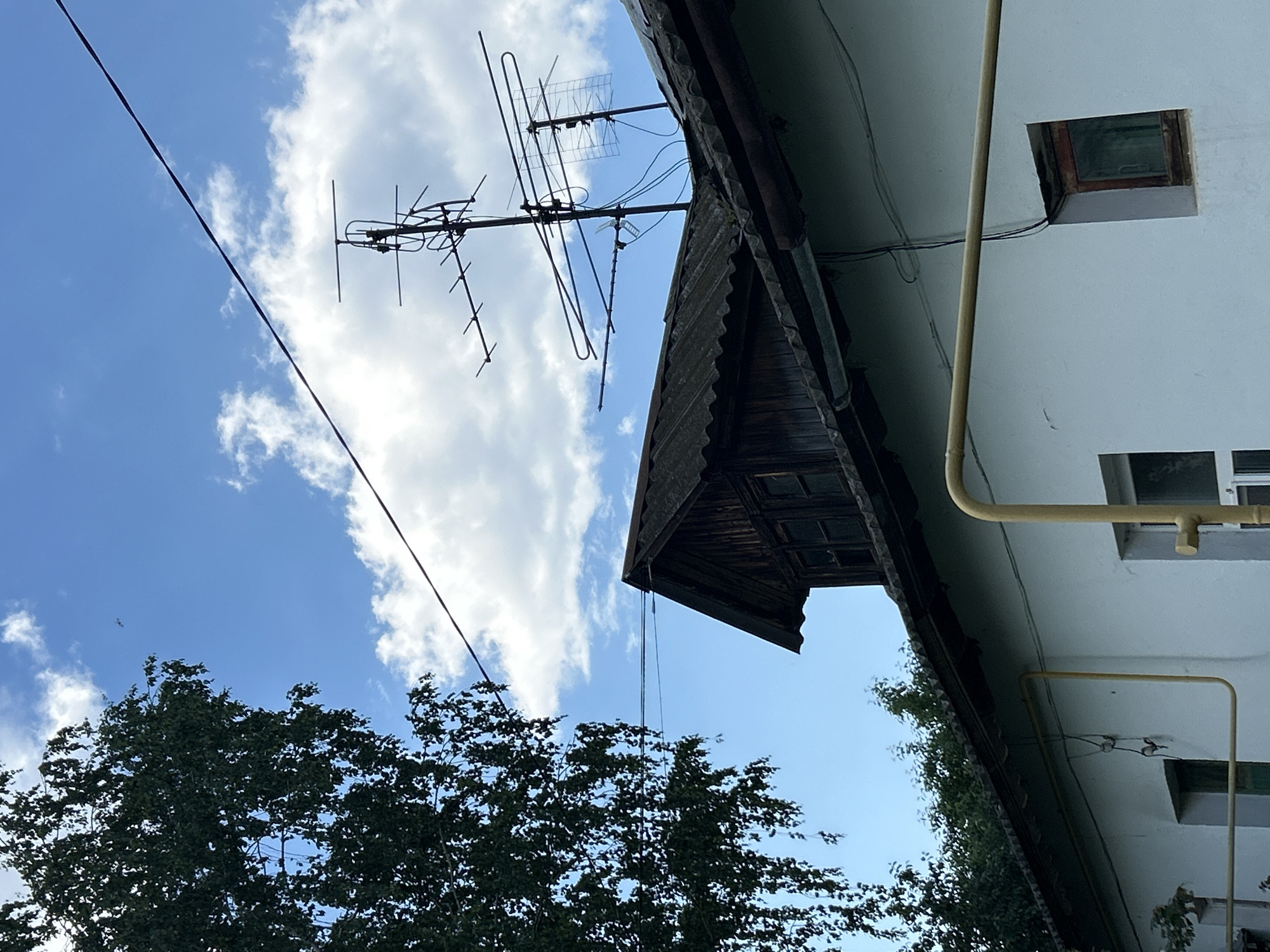 The image captures a segment of a building's exterior under a partly cloudy sky in Chaikovsky, Russia. A large, leafy tree with hanging branches dominates the left side of the frame, its dark green foliage contrasting with the bright blue sky and white clouds. Two wires extend diagonally across the sky, one thick and dark, the other thinner and lighter.
On the roof of the building, an old, multi-element television antenna is prominently displayed. The roof itself appears to be made of dark, corrugated material, and a section of a wooden attic or upper floor is visible, featuring small, dark-paned windows. The building's exterior wall is painted a light, off-white color. A thick, yellow pipe runs horizontally across this wall, with a thinner, darker pipe connected to it and extending downwards. Two windows are visible on the wall; one is partially obscured by the yellow pipe, and the other is a small, square window with a wooden frame. The overall impression is of a somewhat aged residential or utility building in a semi-rural or older urban setting.