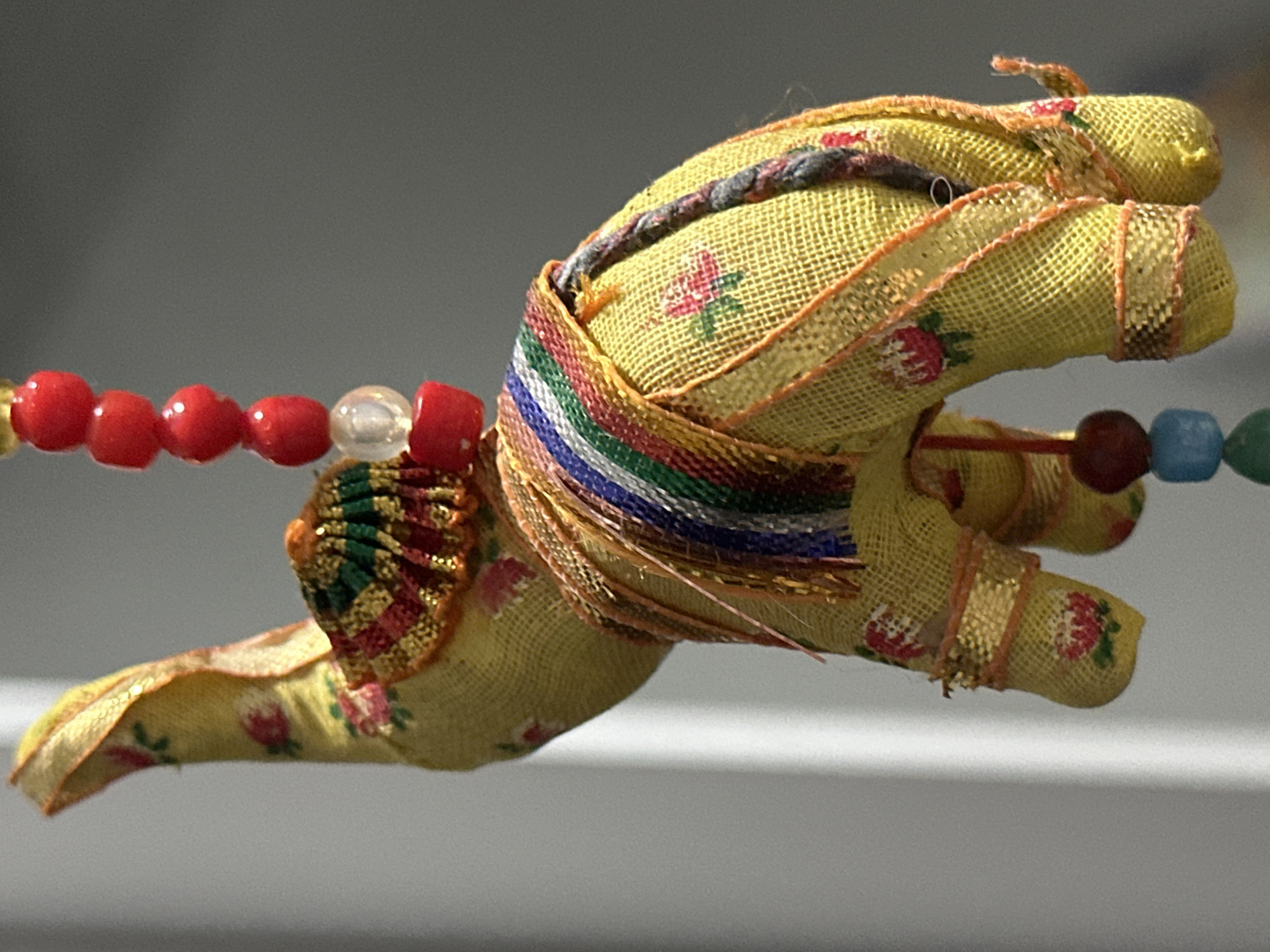This is a close-up, eye-level, medium shot of a decorative stuffed elephant hanging by a string. The elephant is yellow and made of fabric with a floral pattern. It is adorned with colorful trim and beads. The background is out of focus and appears to be a gray wall with a vertical white object casting a shadow. There are no people or animals present in the image. The location is not identifiable within the city of Nevinnomyssk, Russia.