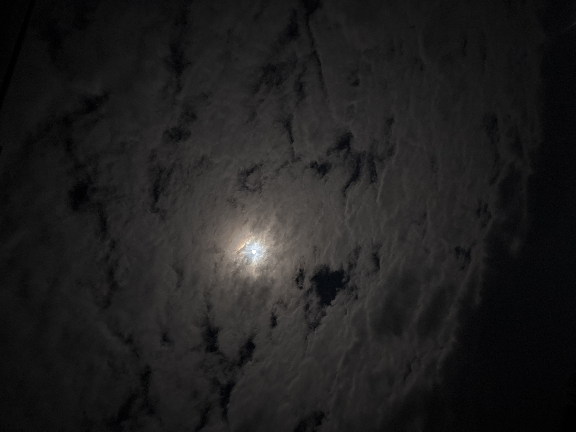 The image shows a dark, cloudy night sky. A bright light, likely the moon, is visible through a break in the cloud cover at the center of the image. The location cannot be determined to be Zlatograd, Bulgaria from the image alone. No people, subjects, objects, actions, or interactions are visible other than the sky. The environment is a night sky with cloud cover and a light source.
