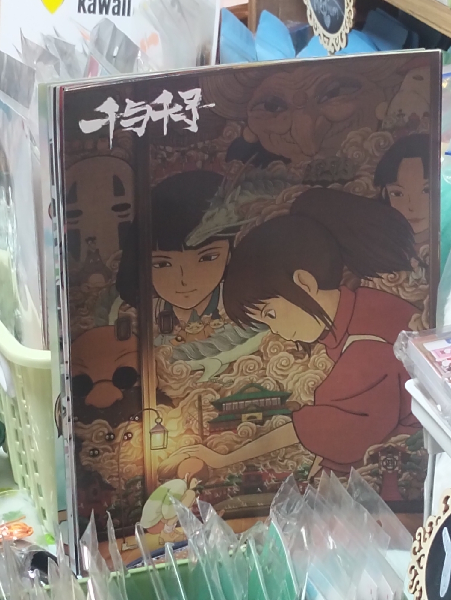 The image displays a close-up view of a framed poster featuring artwork from the animated film "Spirited Away." The poster is placed in what appears to be a retail setting, possibly a store or market stall in Dhaka, Bangladesh, given the context of its presentation within plastic sleeves and the surrounding merchandise.
The artwork on the poster is highly detailed and depicts several key characters and elements from the movie. In the foreground, a young girl with dark hair tied in a ponytail, wearing a red garment, is shown in profile, looking downwards. To her left, another character with dark hair and bangs, facing forward, is partially visible. Dominating the upper portion of the poster is a large, fantastical face with an ornate headdress, resembling a witch or elder figure. Smaller figures, including the masked character No-Face and what appear to be soot sprites, are scattered throughout the composition amidst stylized clouds and architectural elements. A traditional Japanese lantern is also visible, casting a warm glow.
The poster itself is presented upright, leaning against other items. Surrounding the poster, there are glimpses of other merchandise, including more posters or prints in plastic sleeves, suggesting a shop that sells collectible items or art. A light green plastic basket is visible on the left side of the image, partially obscuring the poster. The lighting appears to be ambient, possibly from indoor lighting or a mix of indoor and natural light. The overall impression is that of a display in a commercial establishment.