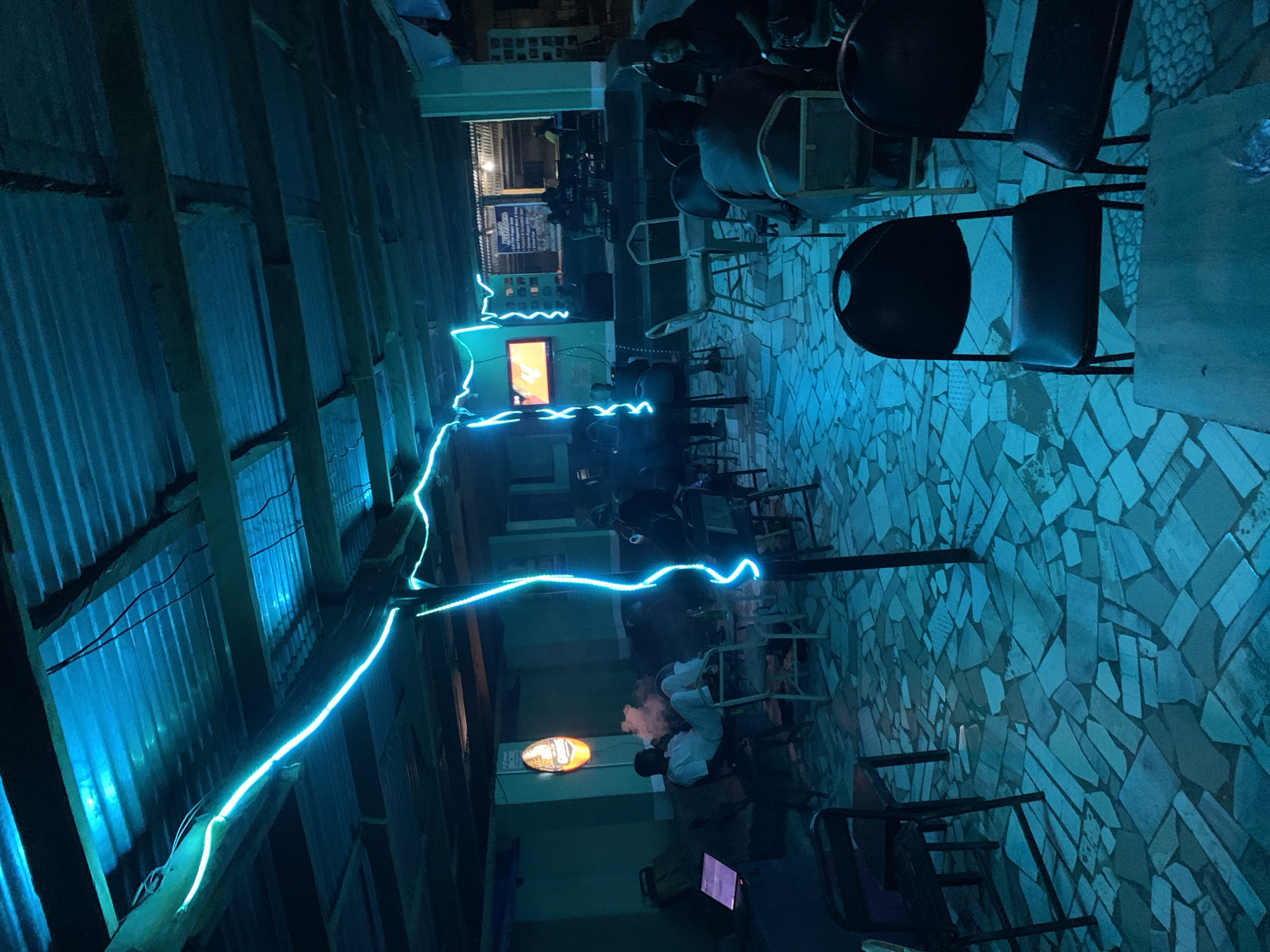 The image depicts an outdoor seating area at night in New Stadium, Nigeria. The area is illuminated with blue light, likely from LED strips, highlighting a metal corrugated roof, wooden supports, and a stone-tiled floor. Numerous black metal chairs and some tables are arranged throughout the space. Several people are present, some seated at tables, suggesting social interaction. A beer sign is visible on a wall. A screen is displaying an image, likely a television screen. Smoke from a cigarette or hookah is visible in the area.