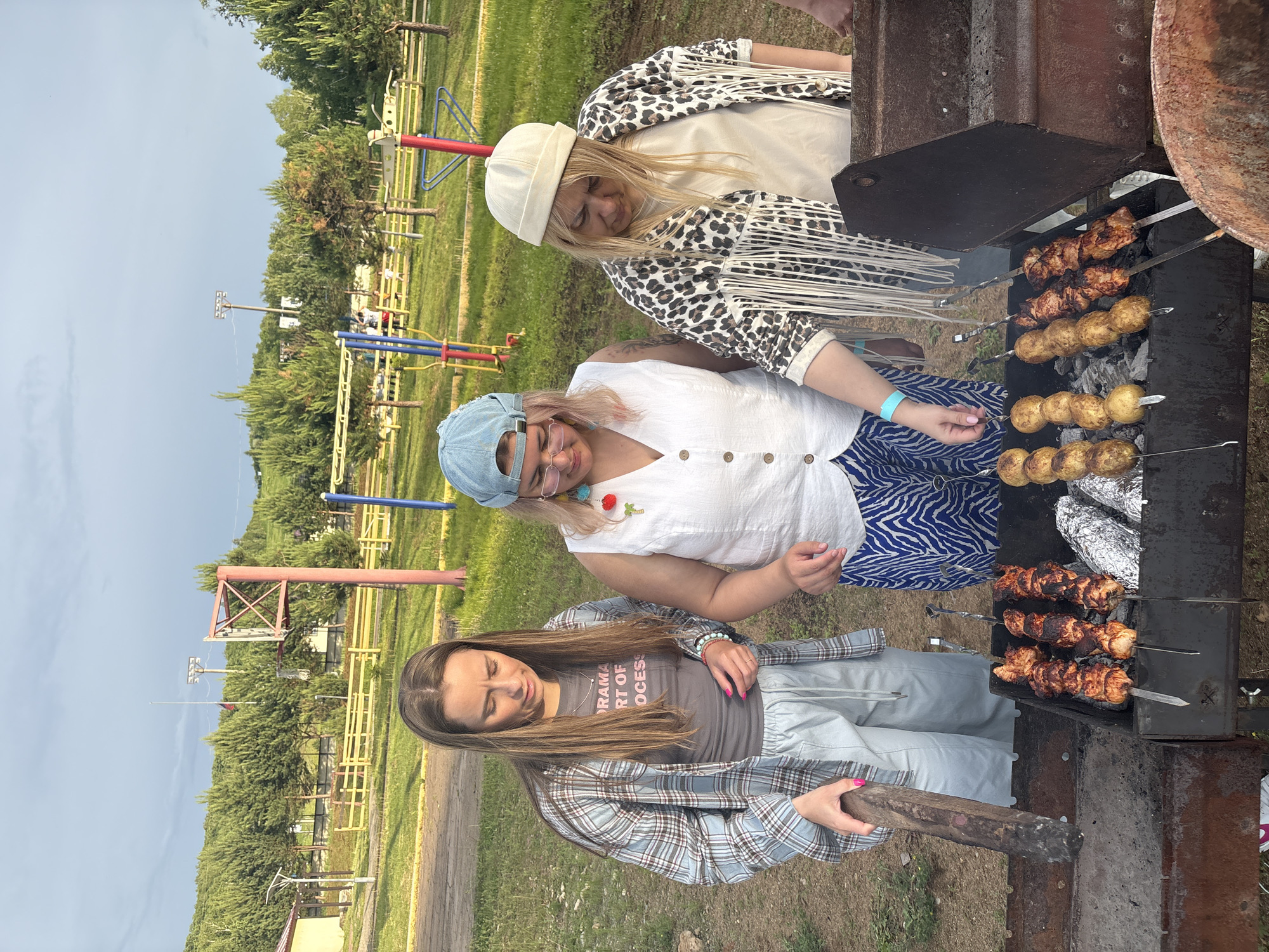 In Birobidzhan, Russia, three women are gathered around a barbecue grill in an outdoor recreational area. The woman on the left, wearing a blue plaid shirt over a grey t-shirt and light blue pants, holds a wooden stick. The woman in the middle wears a denim cap, a white sleeveless shirt, and blue and white striped pants. The woman on the right sports a cream-colored beanie, a cream t-shirt, and a leopard-print jacket with fringe details. They are grilling meat skewers and potatoes. The background features trees, a playground, and a cloudy sky.