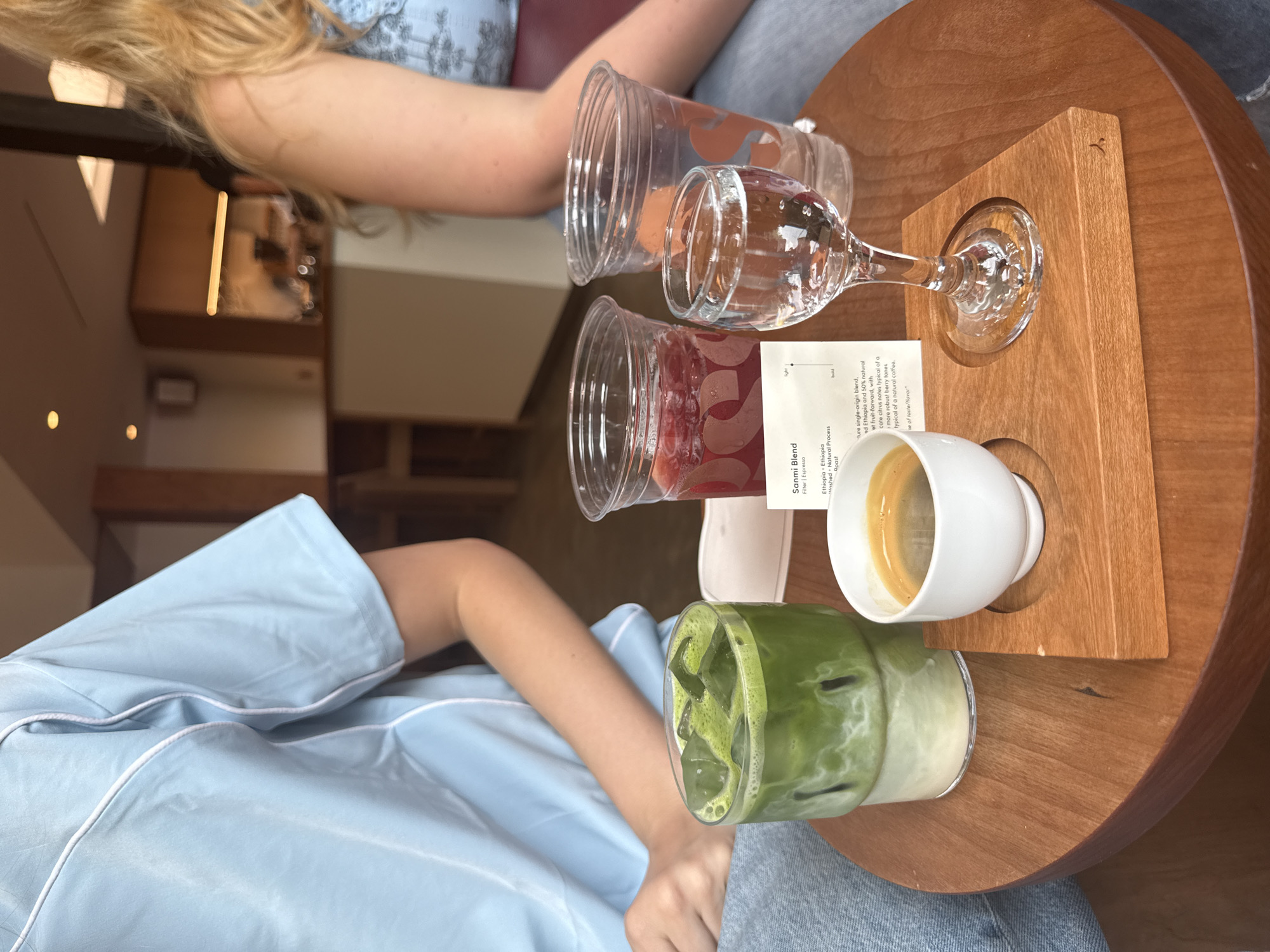 This is a medium shot of a table at a cafe, likely in New York City, United States of America. In the foreground, on a round wooden table, are several drinks. To the left is a tall glass filled with a green matcha latte, layered with white foam and ice cubes. To the right of the matcha latte are two clear plastic cups, one containing a dark red beverage and the other empty. In front of the plastic cups is a small, stemmed glass filled with clear water. Adjacent to the water glass is a small white espresso cup filled with dark espresso, placed on a wooden coaster. A menu card, partially obscured by the water glass, is visible next to the espresso cup. In the background, two individuals are partially visible. One person on the left is wearing a light blue shirt and denim jeans. The other person on the right has blonde hair and is wearing a blue top with a pattern. The cafe environment includes muted lighting and blurred background elements suggesting seating areas and a counter.