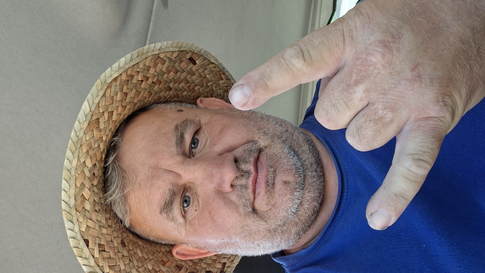 A middle-aged Caucasian man with graying hair and a beard is shown in a close-up, selfie-style shot. He is wearing a straw hat and a blue t-shirt. His right hand is raised, with his index finger and pinky finger extended in a rock-and-roll gesture. His left hand is also visible, with his index finger and thumb extended in a similar gesture. The man appears to be inside a vehicle, with the ceiling of the car visible in the background. The overall lighting suggests an outdoor setting on a bright day. The image was taken on August 1, 2023, in Hîncești, Moldova.