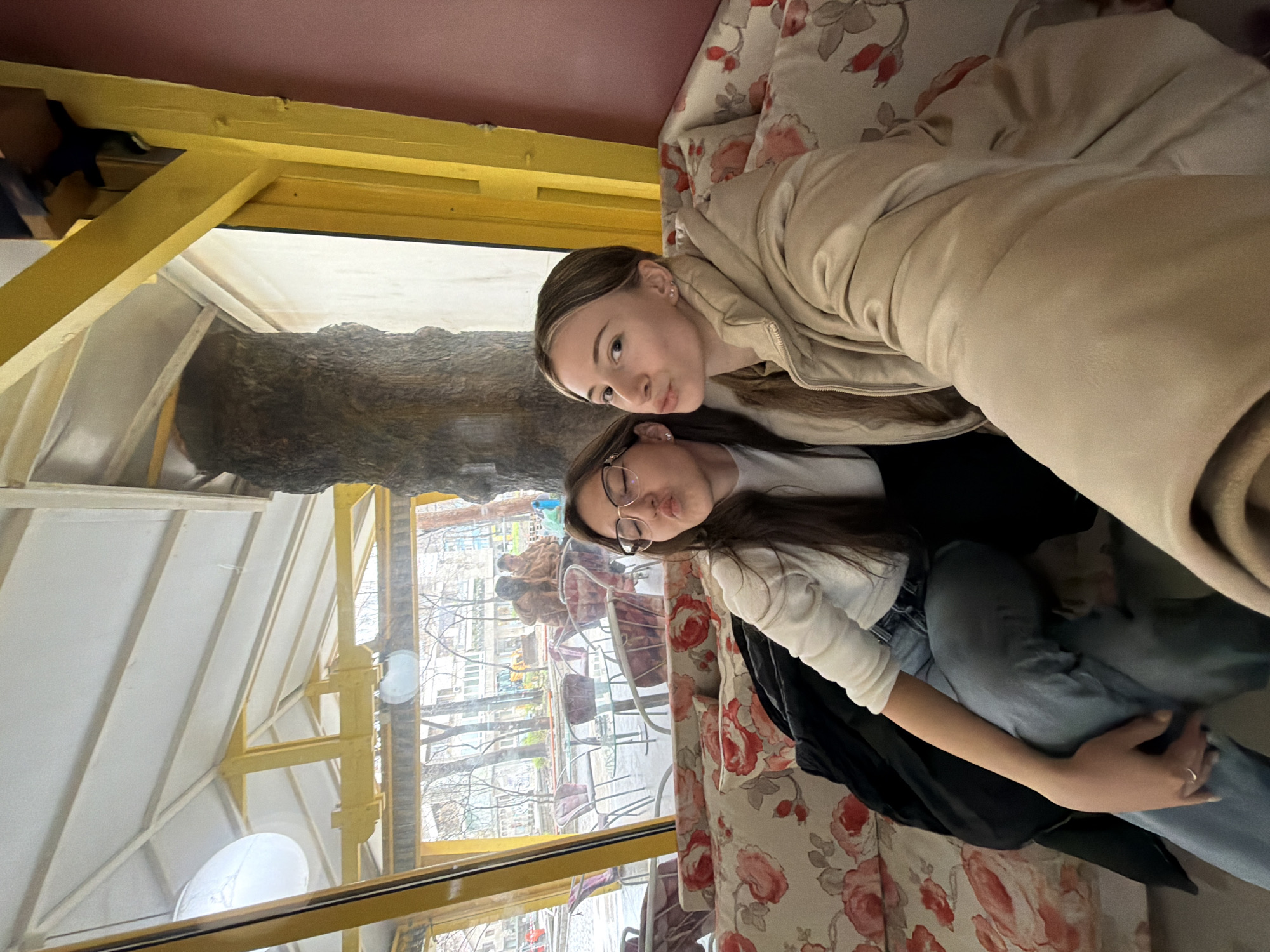 Two young women are posing closely together for a photo inside a restaurant with a unique architectural design. They are likely in a booth with a floral patterned cushion. One of the women wears glasses and is in a white shirt, jeans, and a black jacket. The other woman is wearing a tan jacket. Through the glass window, the architecture of the city of Plovdiv, Bulgaria, can be seen. A large tree trunk is built into the restaurant's interior design. The room has yellow structural beams. Tables and chairs can be seen through the window outside.