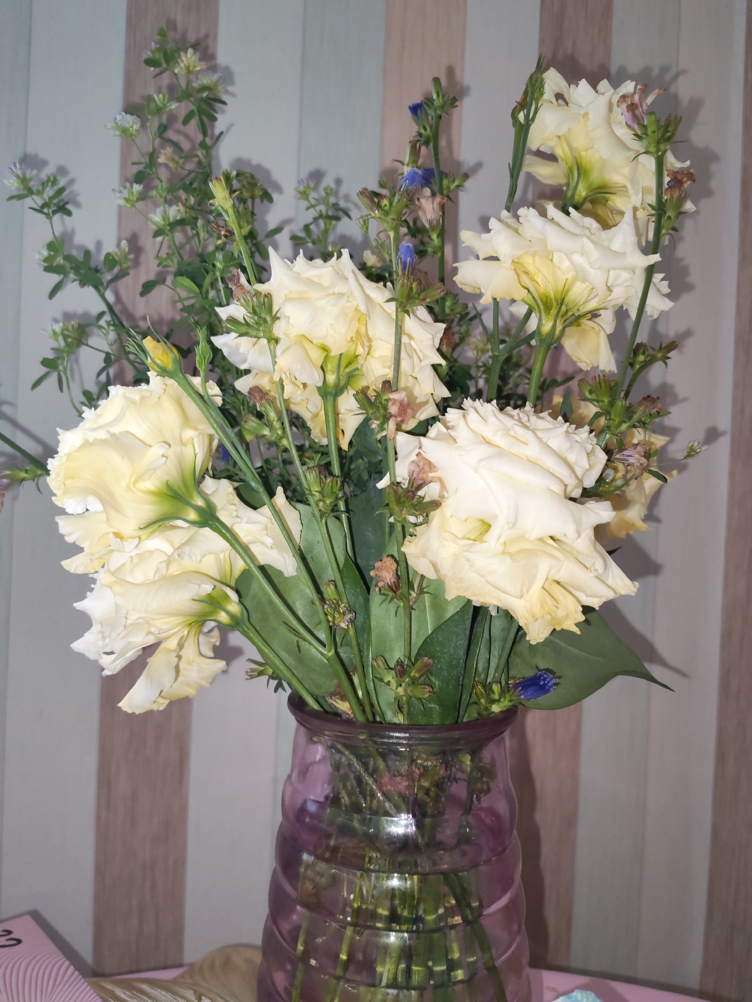 A bouquet of pale yellow flowers with green stems and leaves is displayed in a purple glass vase. The vase has a textured surface with vertical ribs. The bouquet contains several large, open flowers with ruffled petals, along with smaller buds and sprigs of greenery and small blue flowers. The background is a wall with vertical stripes in shades of beige and light gray. A portion of a pink object with wavy lines is visible in the lower left corner. The scene appears to be indoors, possibly in a domestic setting. There are no people or animals present in the image. The lighting suggests an artificial light source. The overall impression is that of a still life arrangement. The location is Izhevsk, Russia.