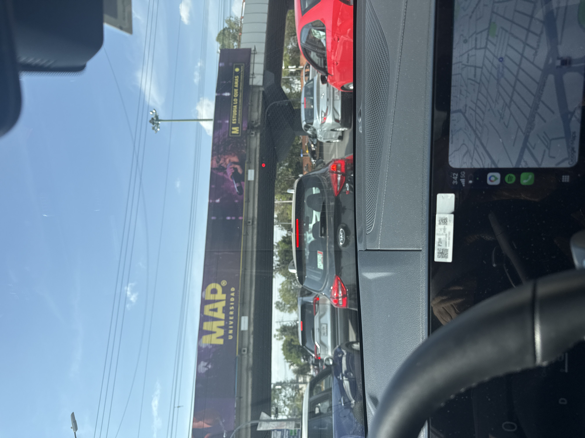 The image, captured from the interior of a car, depicts a street scene with traffic under a bright, clear sky. The view is through the windshield, with parts of the car's dashboard and the steering wheel visible in the foreground.
On the right side of the frame, a digital display in the car shows a map application, likely Google Maps, with street layouts. The screen indicates the time as "3:42" and a "5G" cellular signal. Below the map, icons for Spotify and a phone application are visible. A small white sticker with "P90" and a QR code is affixed near the display.
Outside the car, several vehicles are observed in heavy, possibly stopped, traffic. A red hatchback/sedan is prominently positioned towards the right, followed by a silver car. A dark grey/black Kia SUV, identifiable by its logo and red taillights, is further back in the lane. Other vehicles extend into the distance, suggesting a busy roadway.
In the background, a large billboard stretches across the scene. It features text in yellow and white, reading "M ESTUDIA LO QUE AMAS" (M STUDY WHAT YOU LOVE) and "MAP. UNIVERSIDAD". Above the traffic, an elevated concrete structure, possibly an overpass or bridge, is present. The sky is a vibrant blue with sparse white clouds, indicating sunny daytime conditions. Utility lines crisscross the upper part of the frame, and a tall street light fixture is also visible. The scene portrays an urban or suburban environment, consistent with the provided location context of Puebla, Mexico.