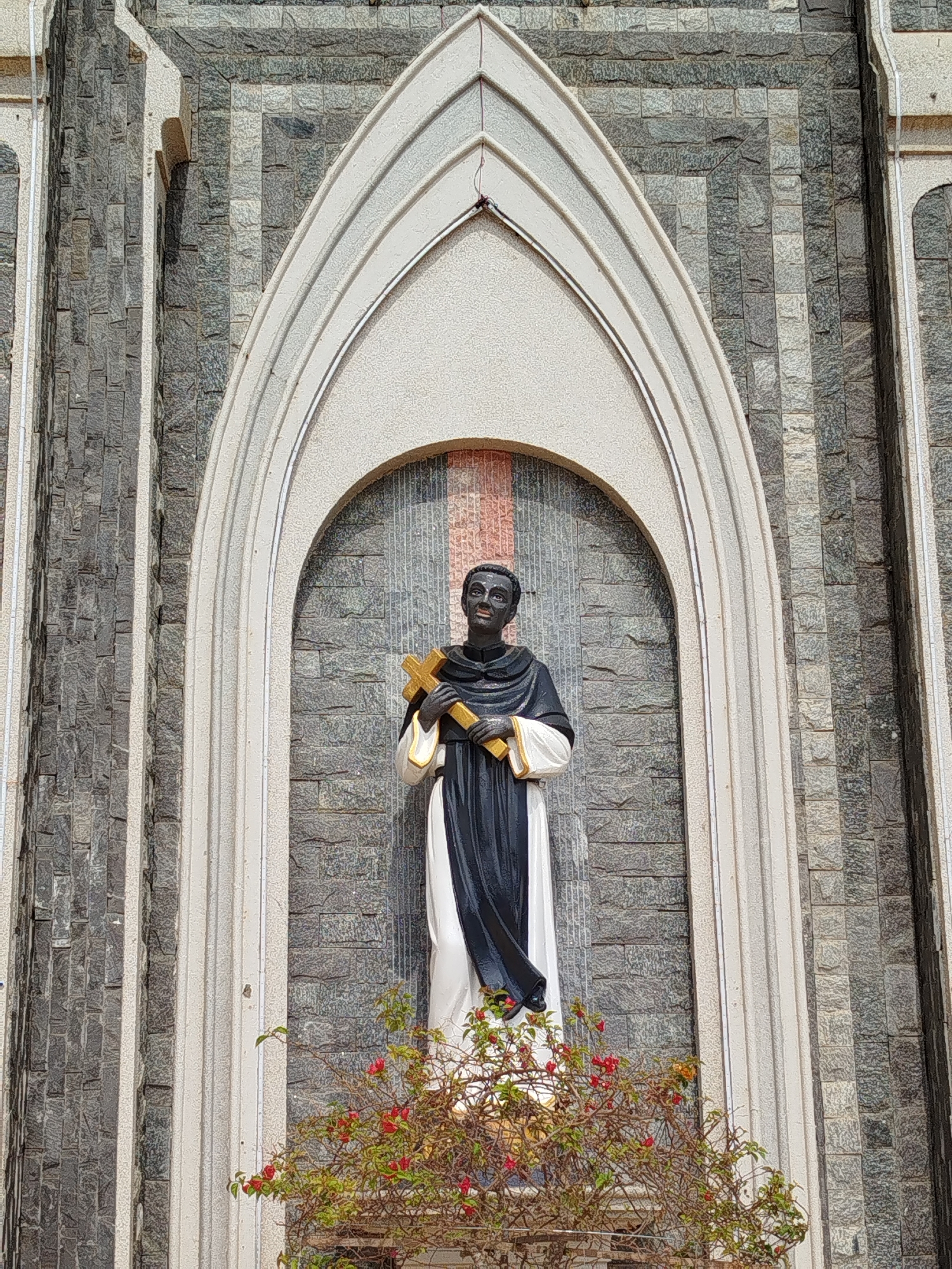 The image captures a full-length statue of a male religious figure, likely a saint, positioned centrally within an elaborate arched niche on the facade of a building. The statue depicts a dark-skinned man wearing a black cloak over a white robe, holding a prominent golden cross against his chest with both hands. His gaze is directed slightly upwards, conveying a solemn or contemplative expression.
The arched niche is framed by multiple layers of light-colored, possibly concrete or stone, moldings that contrast with the darker background wall. This wall is constructed from stacked rectangular blocks of grey and dark grey stone, creating a textured pattern across the building's surface. To either side of the central niche, further vertical architectural elements, matching the light-colored material of the arch, are visible.
At the base of the statue, a small, leafy bush with bright red flowers provides a splash of natural color. The scene is brightly and evenly lit, suggesting daytime conditions. No discernible text is visible within the frame. The architecture indicates a religious building, possibly a church or cathedral, located in Giãn Dân, Vietnam.