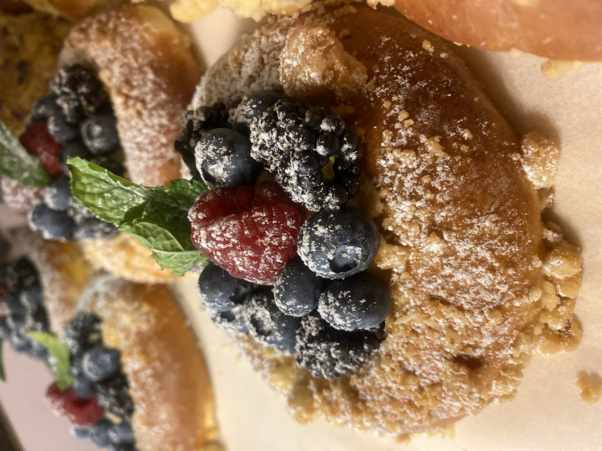 A close-up shot shows a pastry topped with fresh berries and powdered sugar. The pastry itself has a golden-brown crust and appears to be sprinkled with small, crumbly pieces. Berries, including blueberries and raspberries, are arranged on top, with a sprig of mint providing a green accent. The pastry is dusted with powdered sugar, creating a delicate white coating. The background is out of focus, suggesting the pastry is part of a larger display or meal. The lighting is soft, highlighting the textures and colors of the food. No information about location in Warsaw, Poland is available in the image.