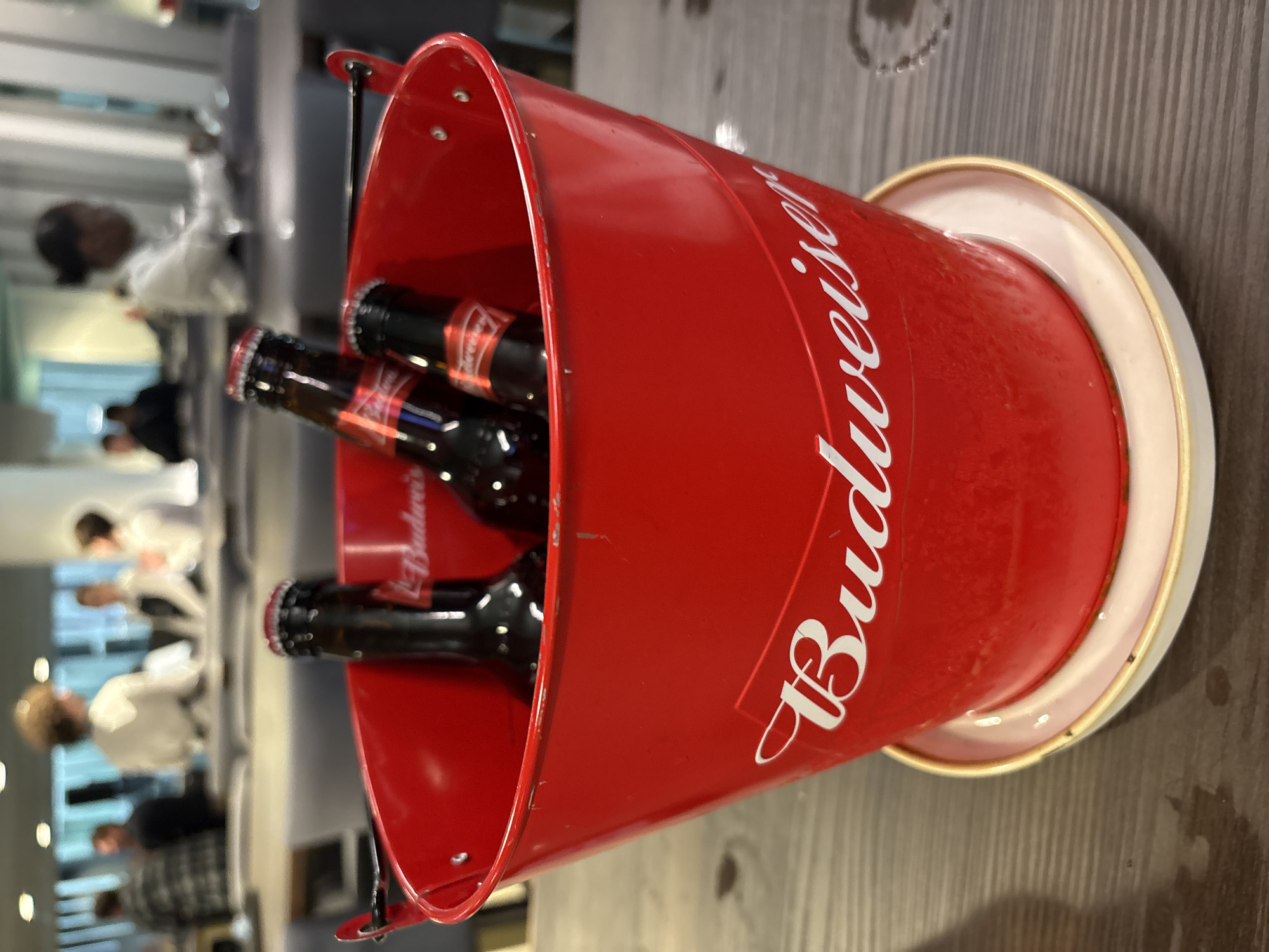 The image depicts a red Budweiser-branded bucket filled with bottles of Budweiser beer. The bucket sits on a wooden surface. In the background, blurred figures suggest a public space or restaurant setting, potentially in London, United Kingdom. There are individuals sitting or standing. The lighting appears to be interior. The primary focus is on the beer bucket and its contents.
